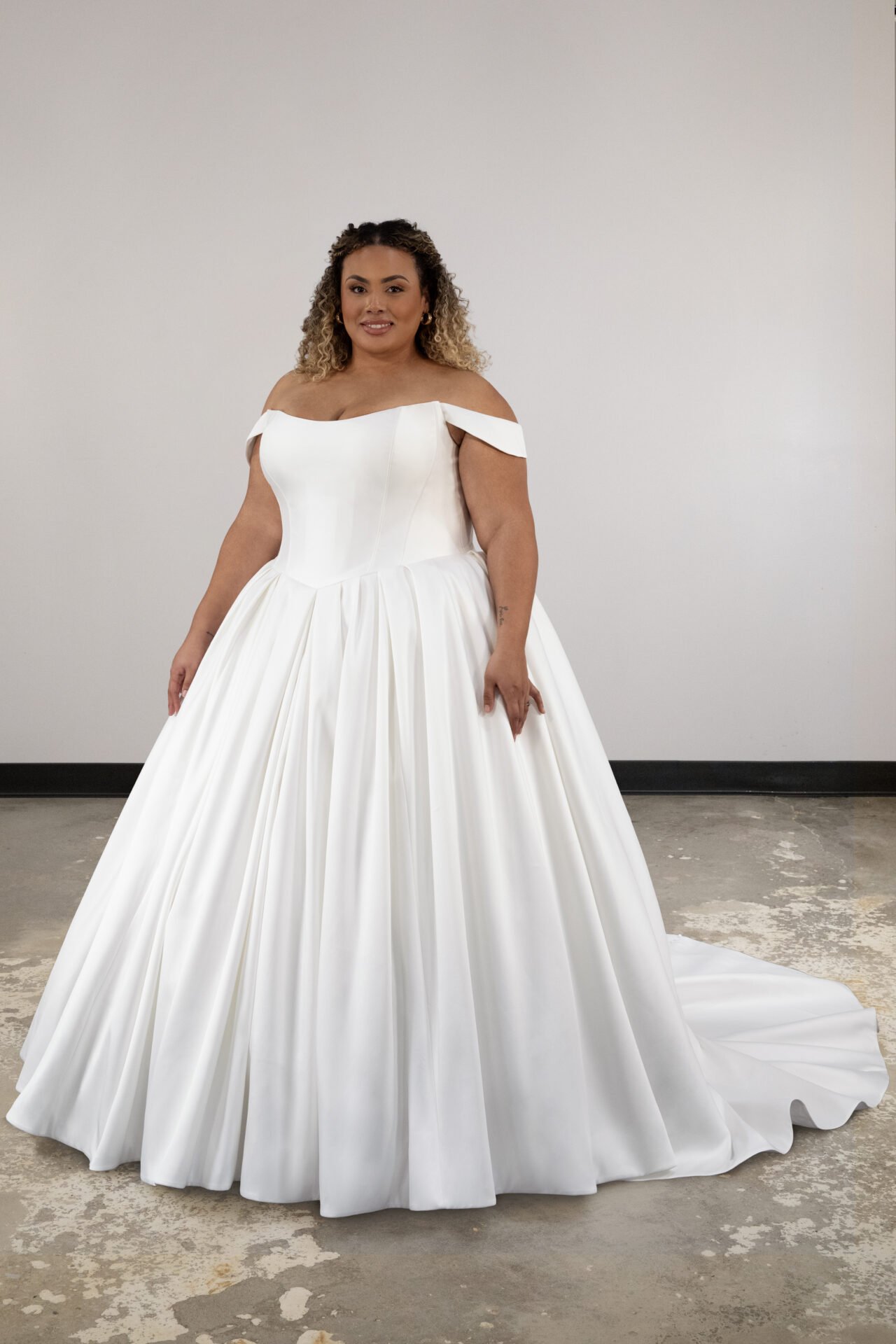 Woman wearing an off-the-shoulder white wedding gown with a full skirt, standing against a plain background.