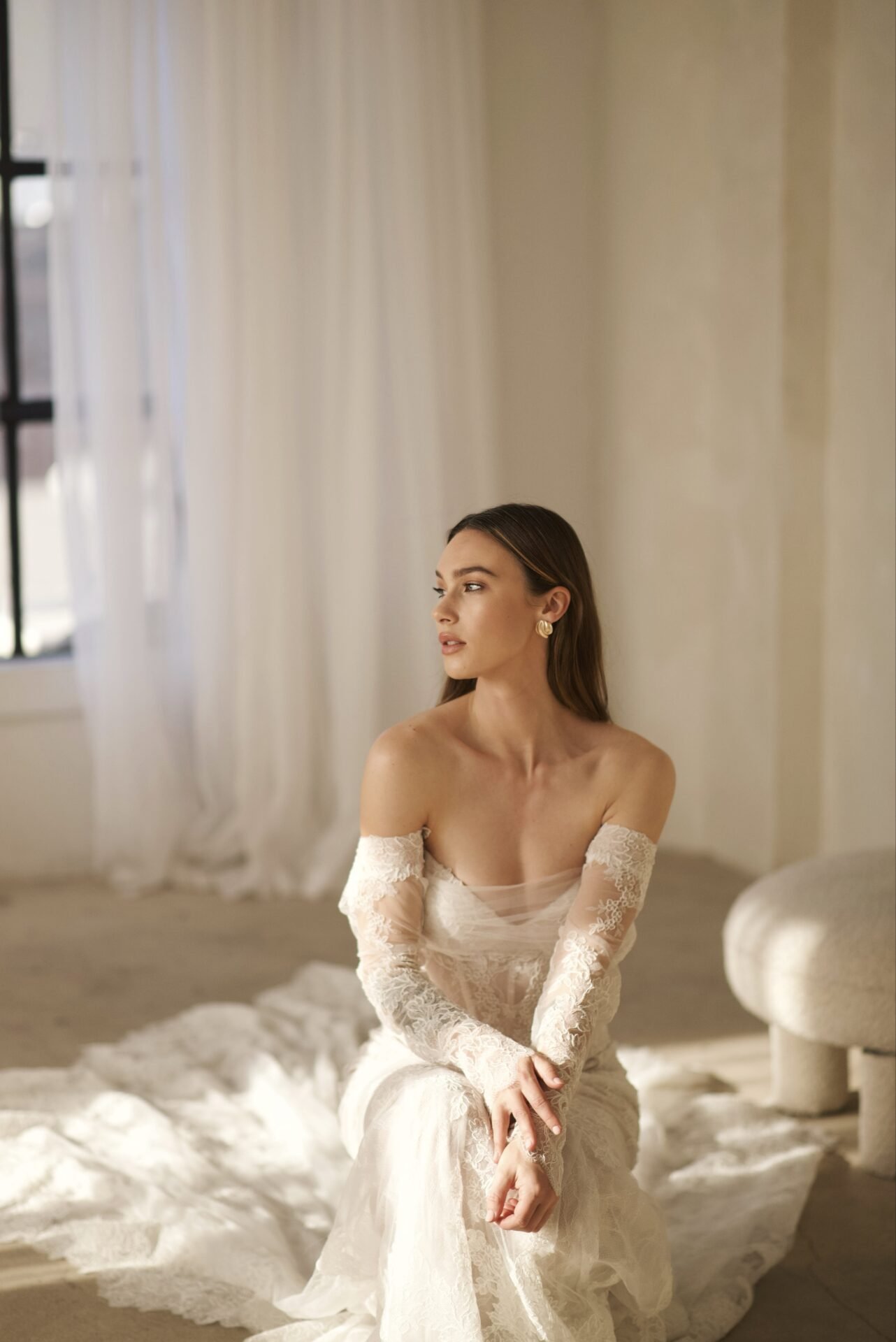 A woman in an off-shoulder lace wedding dress sitting on a white fluffy rug in a softly lit room with white curtains.