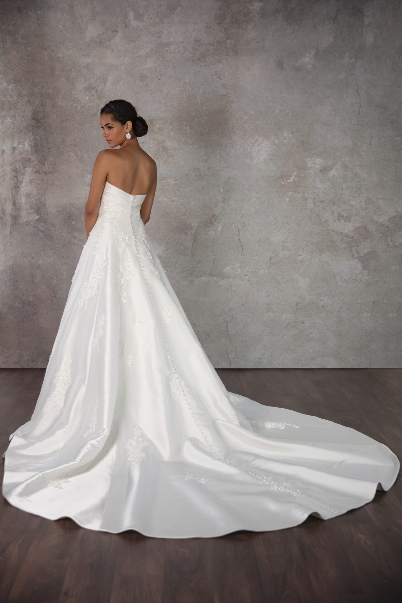 A woman in an elegant white wedding gown with embroidery, standing against a textured gray wall, looking over her shoulder with her hair in a low bun and earrings.