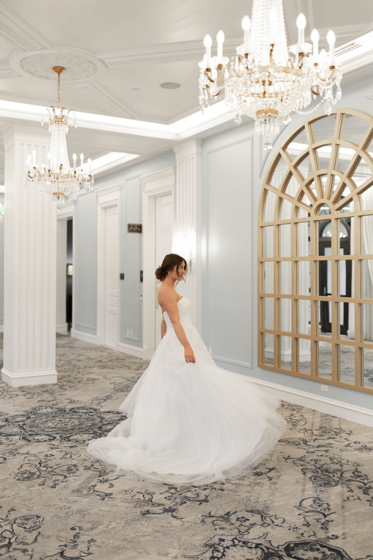 A bride in a white wedding gown standing in an elegant, well-lit room with chandeliers, light-colored walls, and ornate paneling.