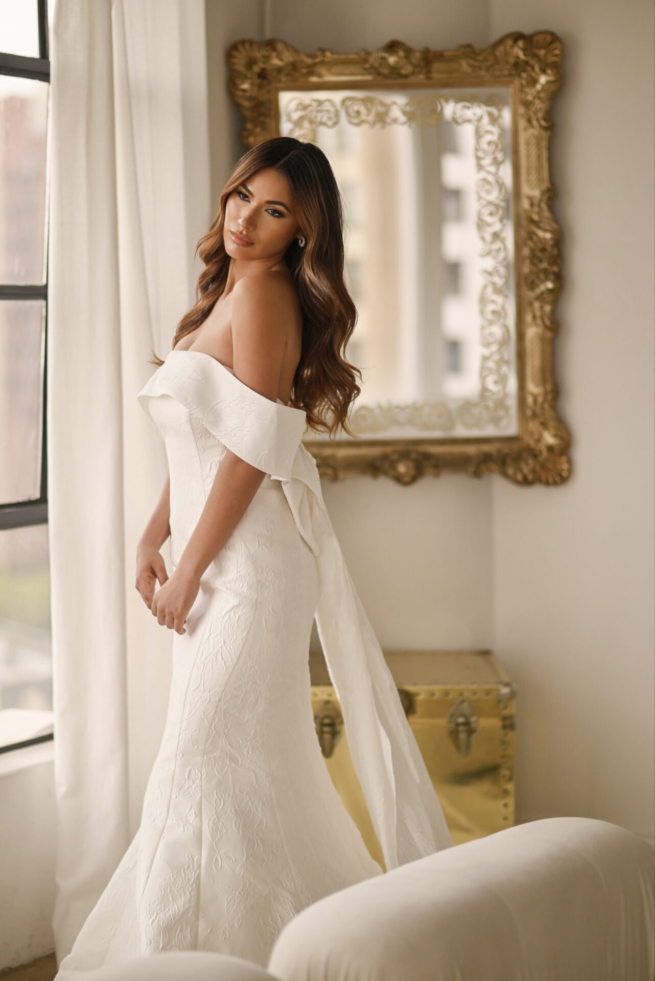 A woman in an off-the-shoulder wedding dress with a bow detail, standing next to a window with white curtains and a large ornate gold mirror behind her.
