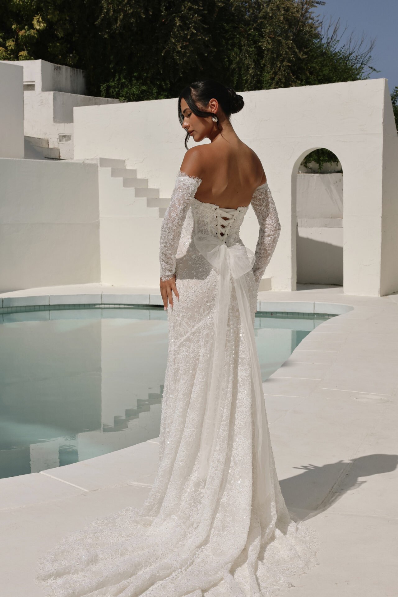 A woman in a white lace wedding dress with off-the-shoulder sleeves standing outdoors near a small pool, with white stucco structures and greenery in the background.
