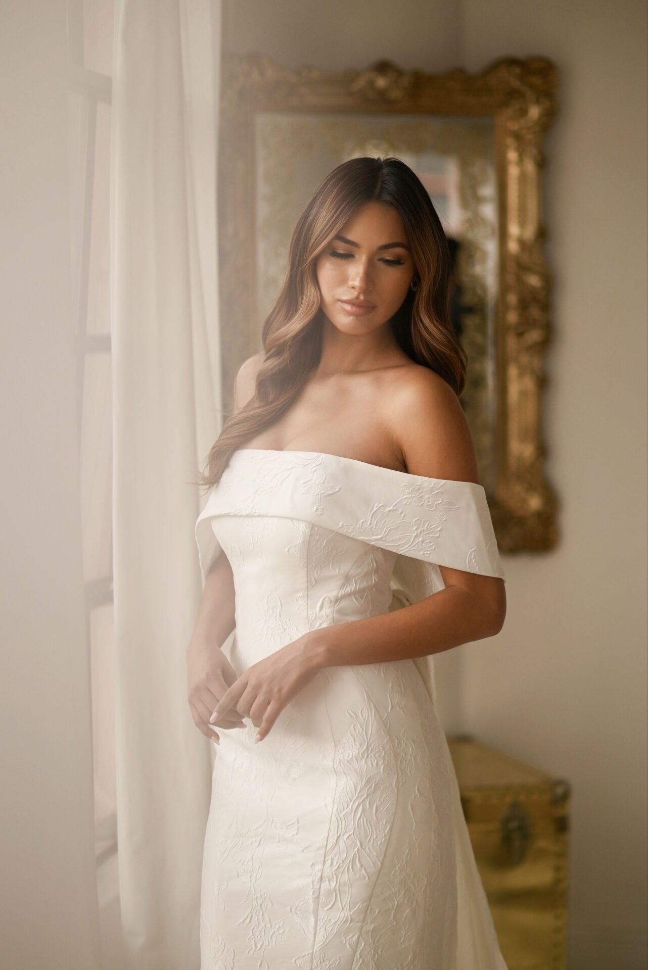A woman in a white off-shoulder wedding dress with embroidered details, standing near a window with sheer curtains, in front of a large ornate gold-framed mirror.