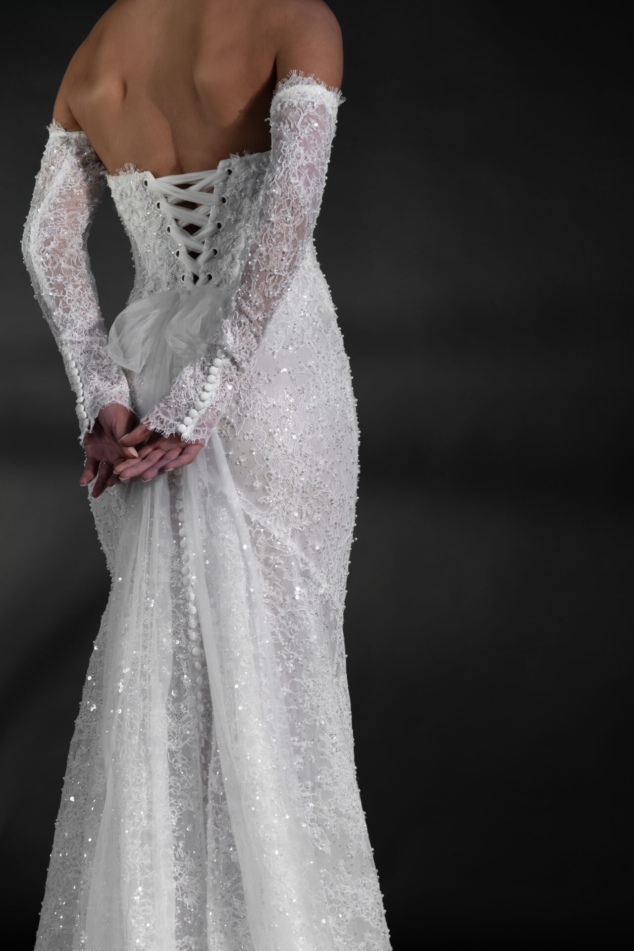 Back view of a person wearing a white lace wedding dress with long sleeves, lace-up back, tulle accents, and intricate beaded detailing against a dark background.