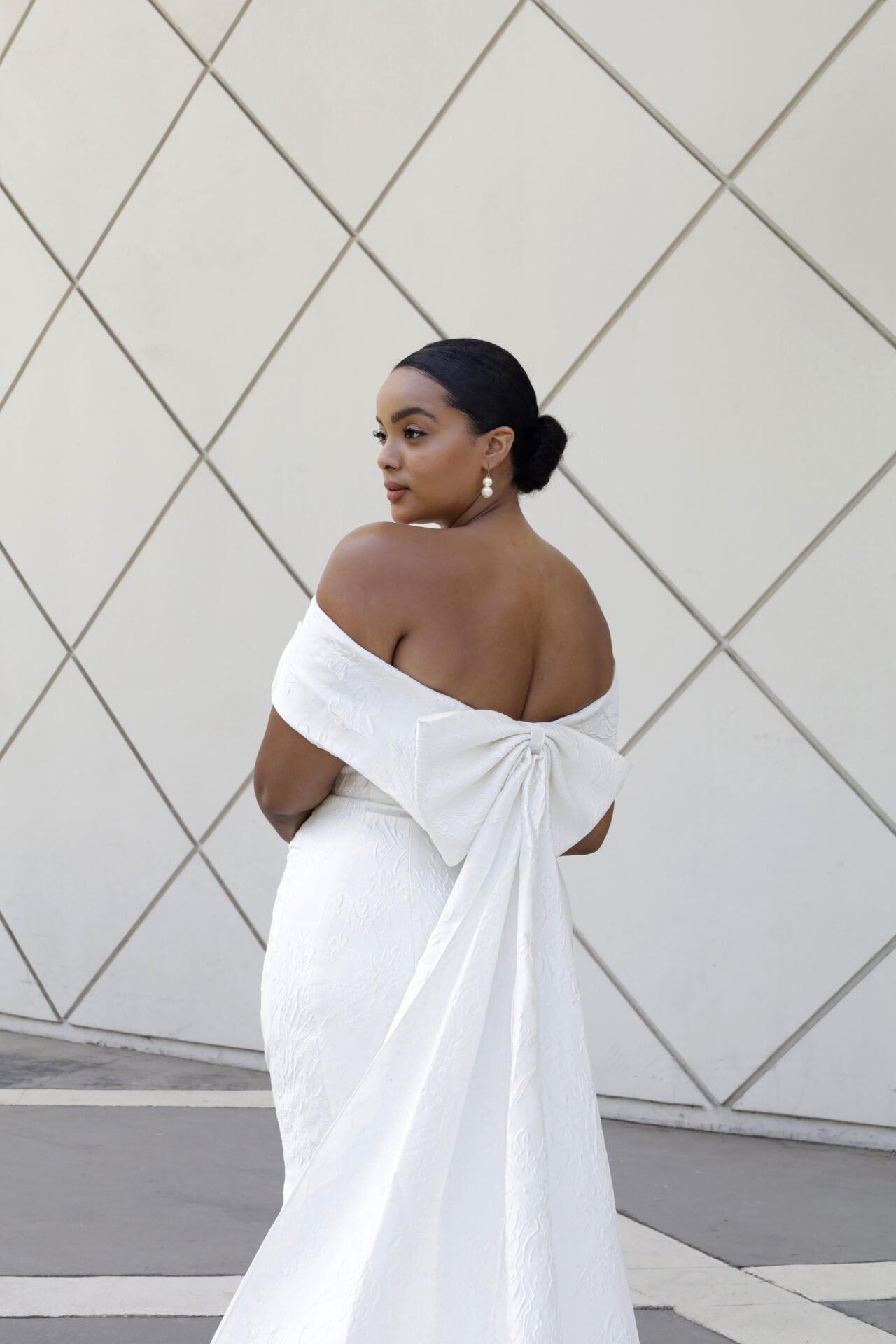 A woman with dark hair in a bun wearing pearl earrings and a white off-shoulder gown with a large bow at the back, standing against a geometric-patterned wall.