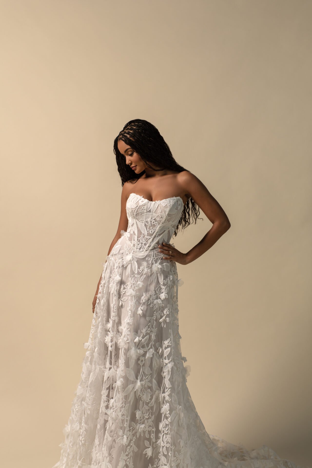A woman in a white, strapless, embroidered wedding dress with floral lace details, standing against a plain beige background.