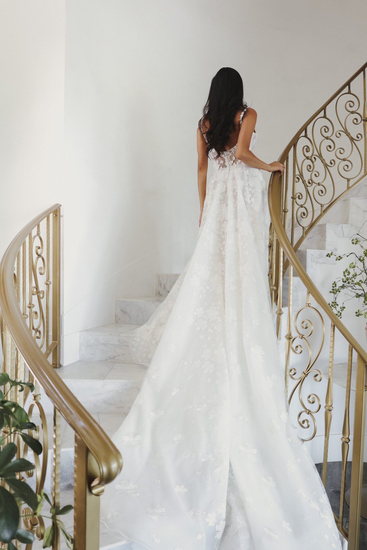 A woman in a wedding dress standing on a marble staircase with gold ornate railing, facing away from the camera.