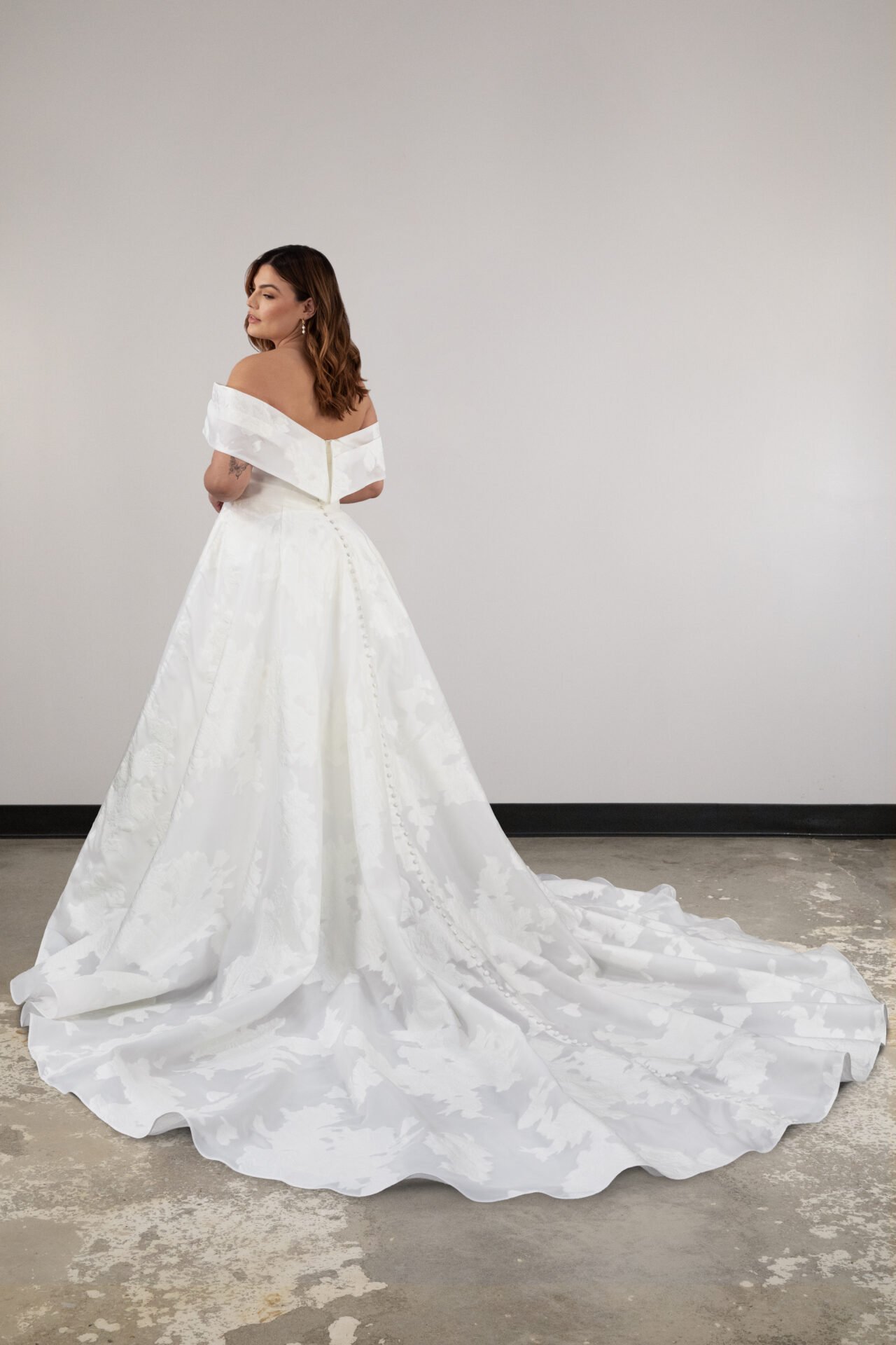 A woman in a white off-shoulder wedding gown with a long train standing against a plain white wall.