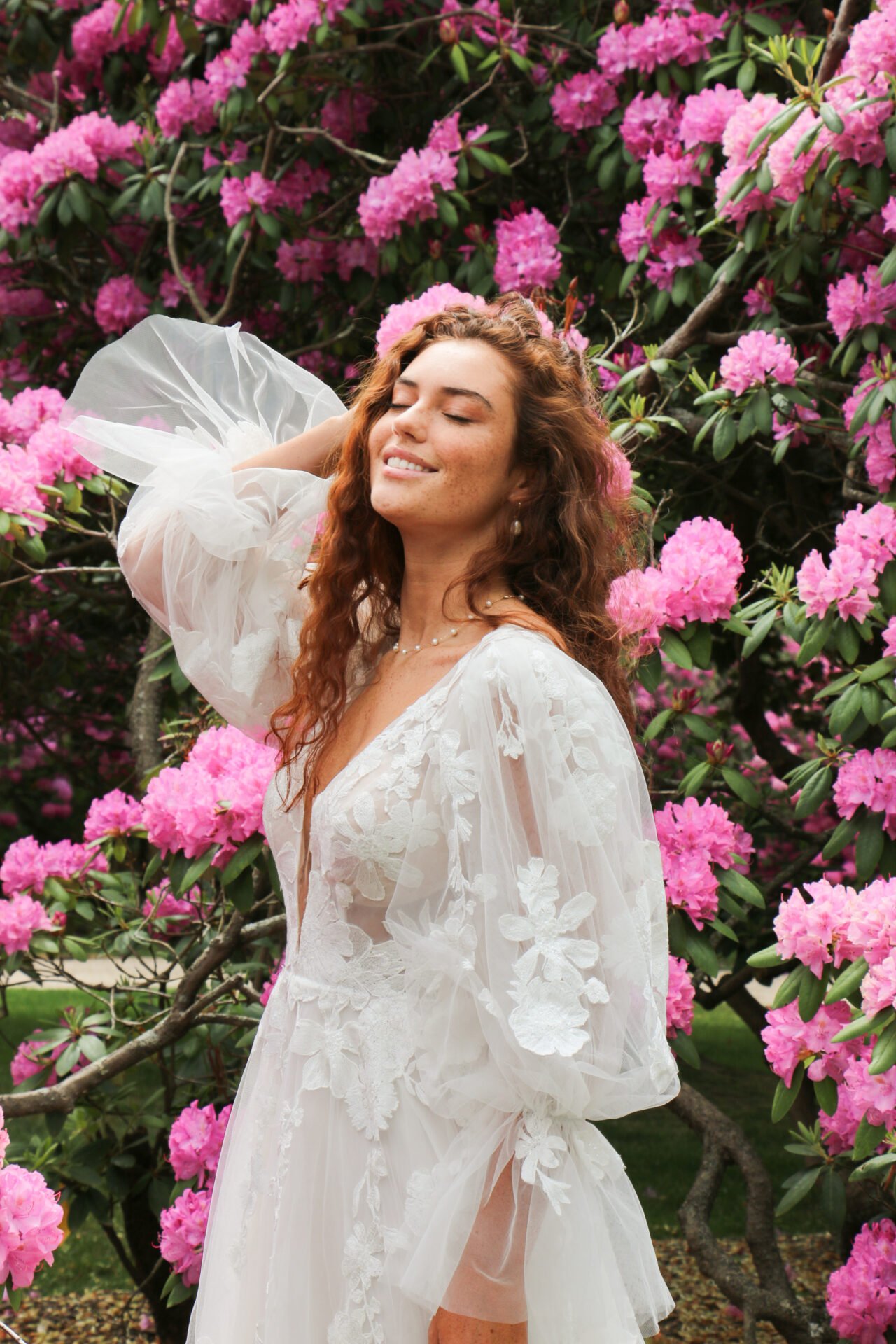 A woman with long curly red hair in a white lace dress standing among pink flowering bushes, smiling with eyes closed.