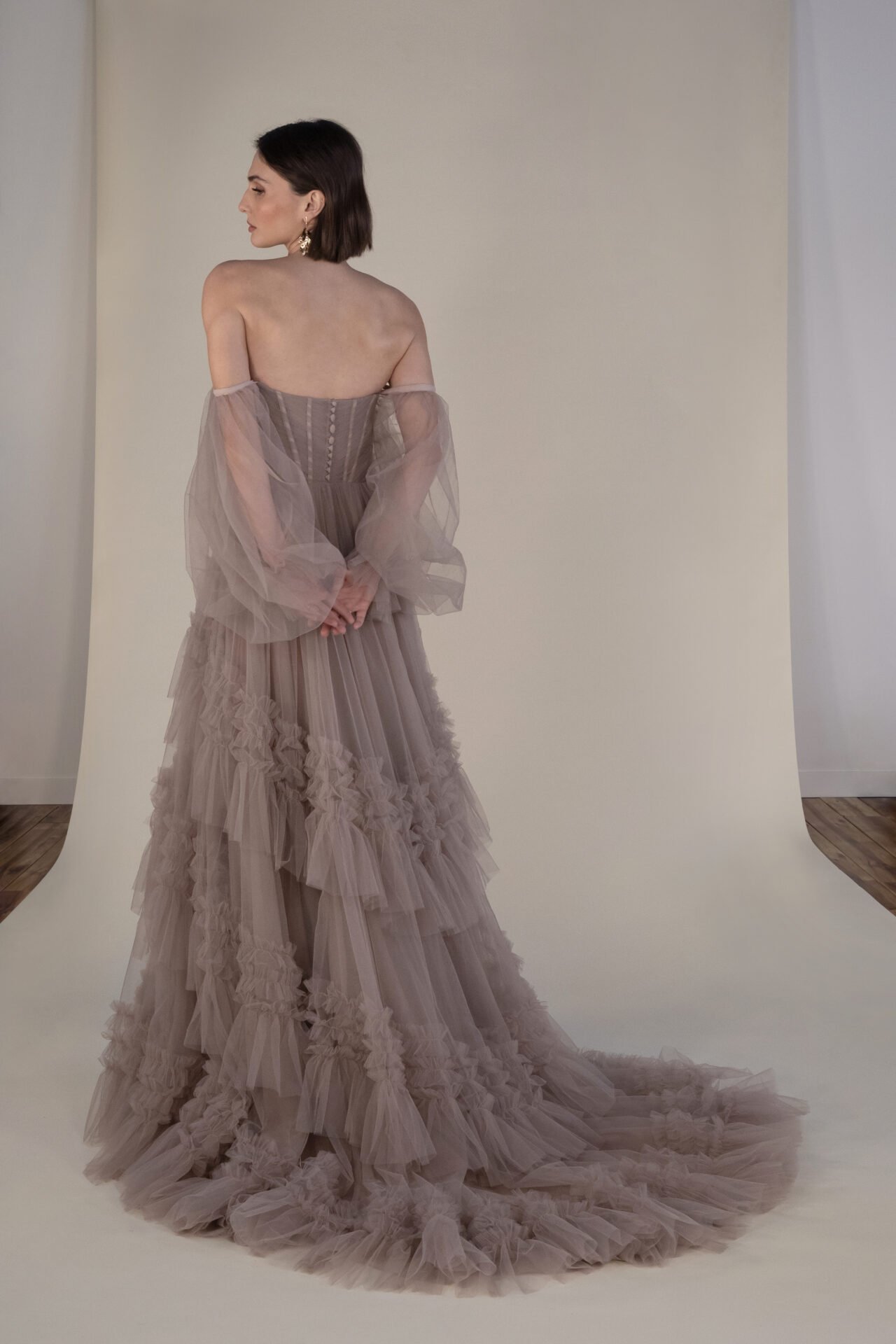 A woman in an elegant, ruffled, beige gown standing with her back to the camera in front of a beige background.