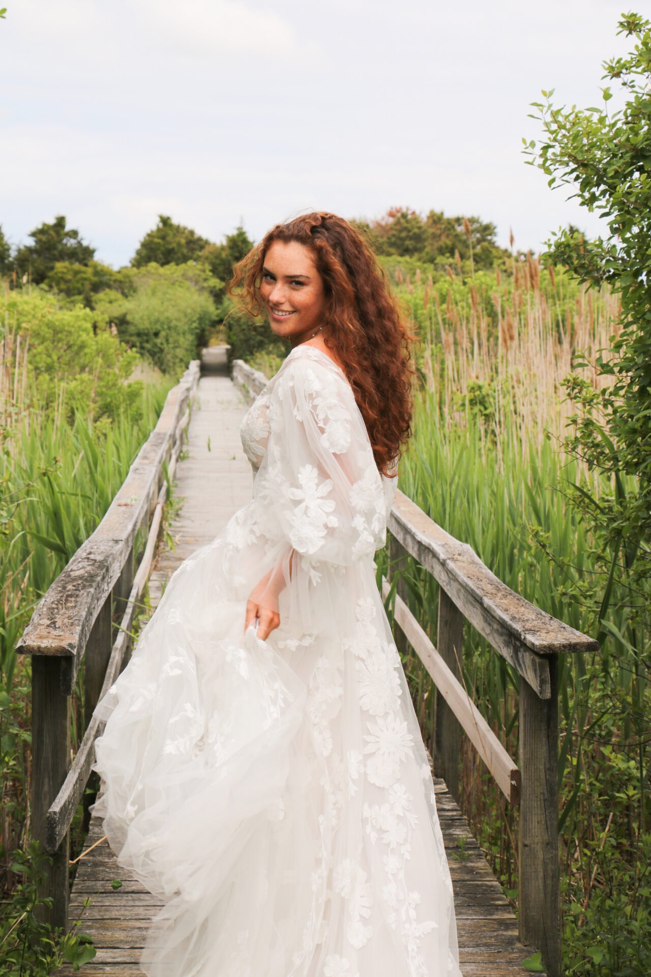 Young woman in a white lace dress standing on a wooden bridge surrounded by green vegetation, smiling and looking back.