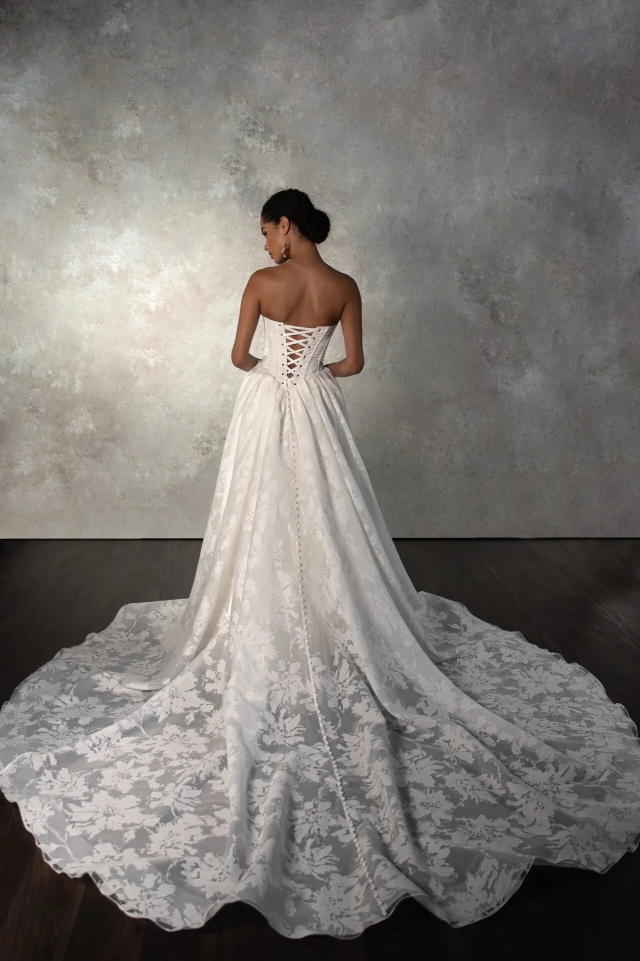 Back view of a woman in a strapless white wedding gown with a long train, standing against a textured gray wall.