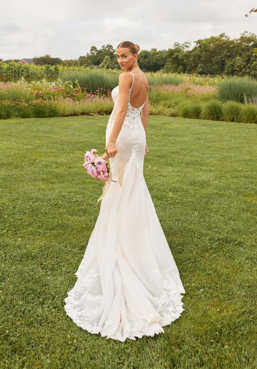 Bridal model in a white lace wedding gown with an open back, holding a bouquet of pink flowers, standing on a green lawn with trees and flowering shrubs in the background.