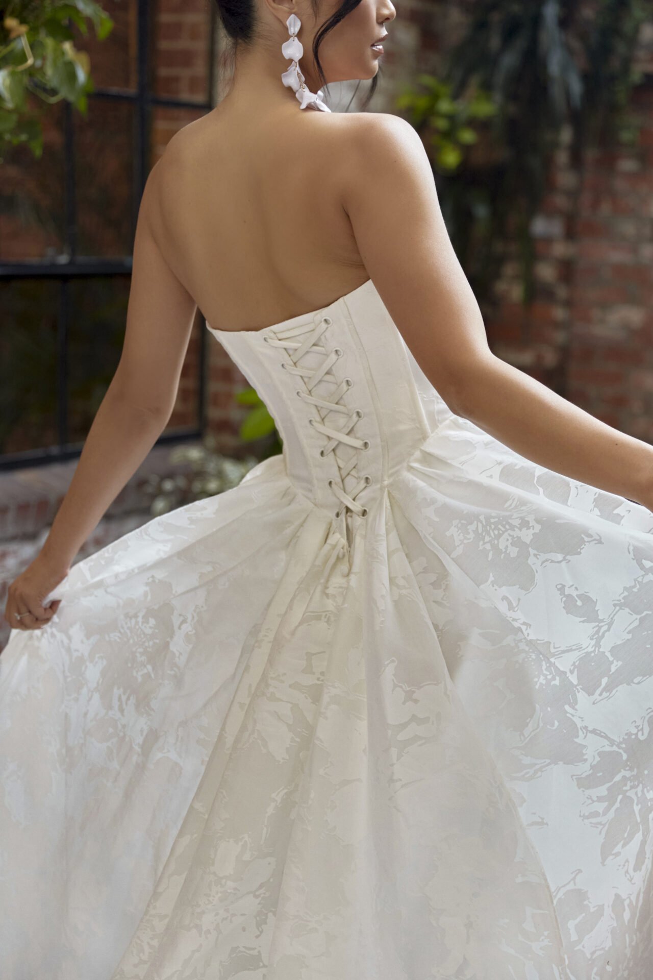 A woman wearing a strapless white wedding dress with laced-up back, holding the skirt, standing indoors near brick walls and plants.