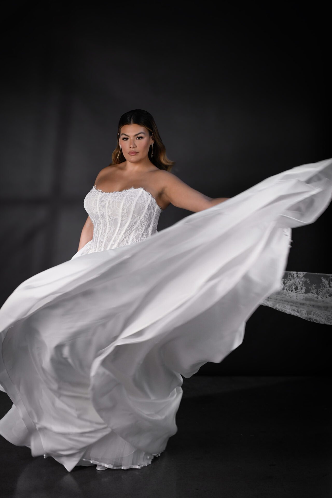 A woman in a white strapless wedding dress with lace details poses against a dark background, holding the flowing fabric of her dress.