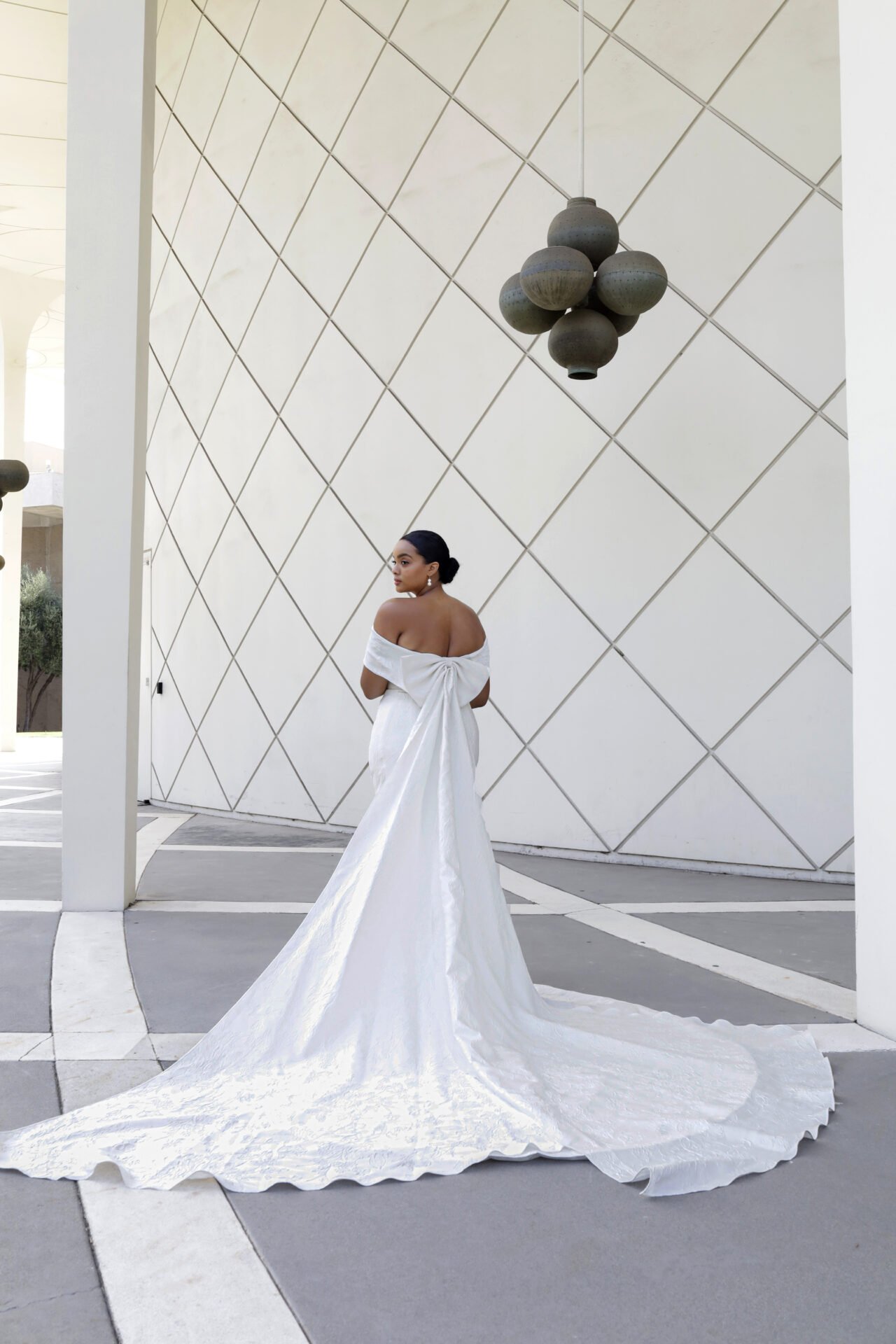 A woman in a white off-shoulder wedding dress with a long train standing against a modern white wall with geometric patterns, outdoors during daytime.