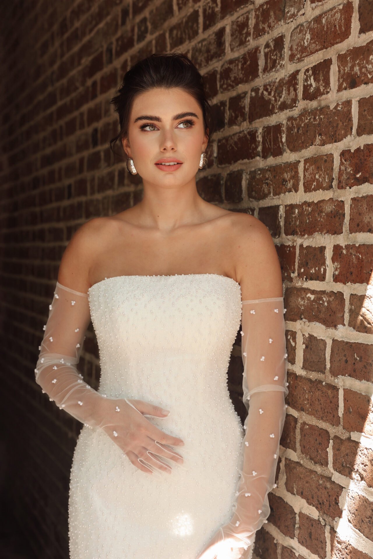 A woman in an elegant off-shoulder wedding dress with sheer, beaded long sleeves, standing against a brick wall.