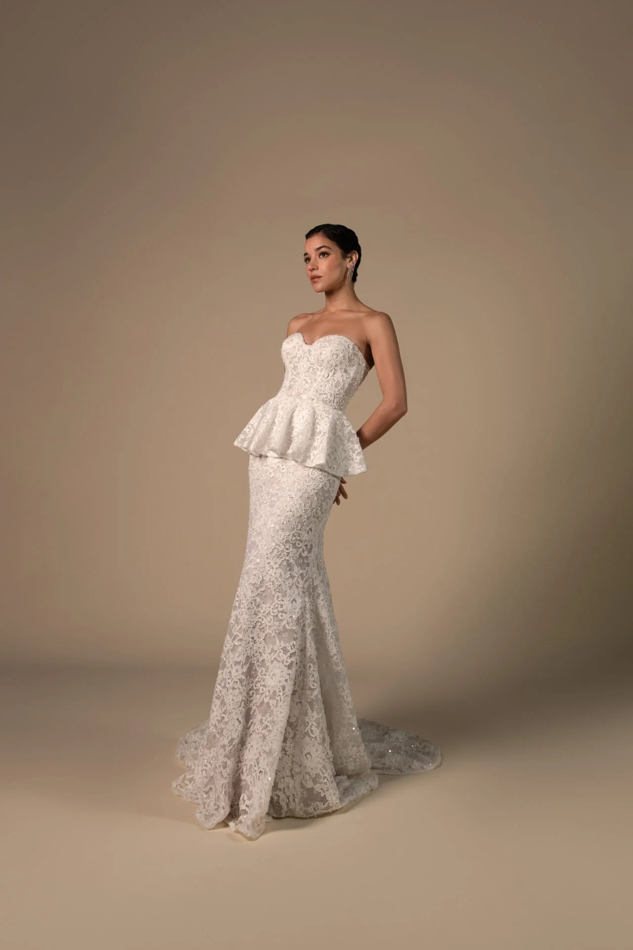 A woman in a white lace wedding gown with a fitted bodice and peplum detail, standing against a plain beige background.