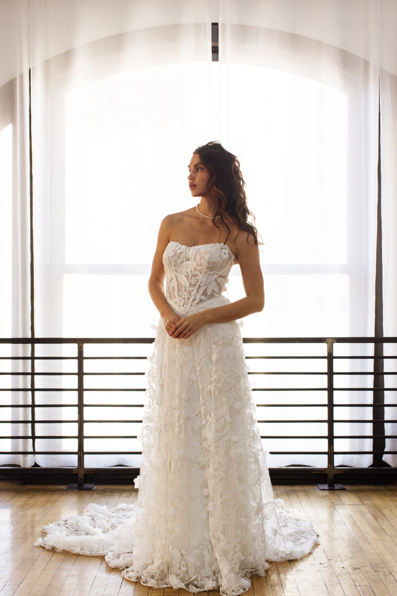 Woman in a white lace wedding dress standing indoors near a large window with sheer curtains, backlit by natural light.