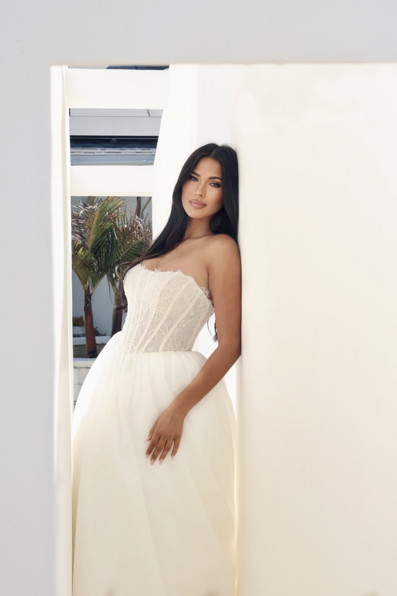 A woman in a white strapless dress with lace bodice and flowing skirt posing against a white wall with a window showing palm trees outside.