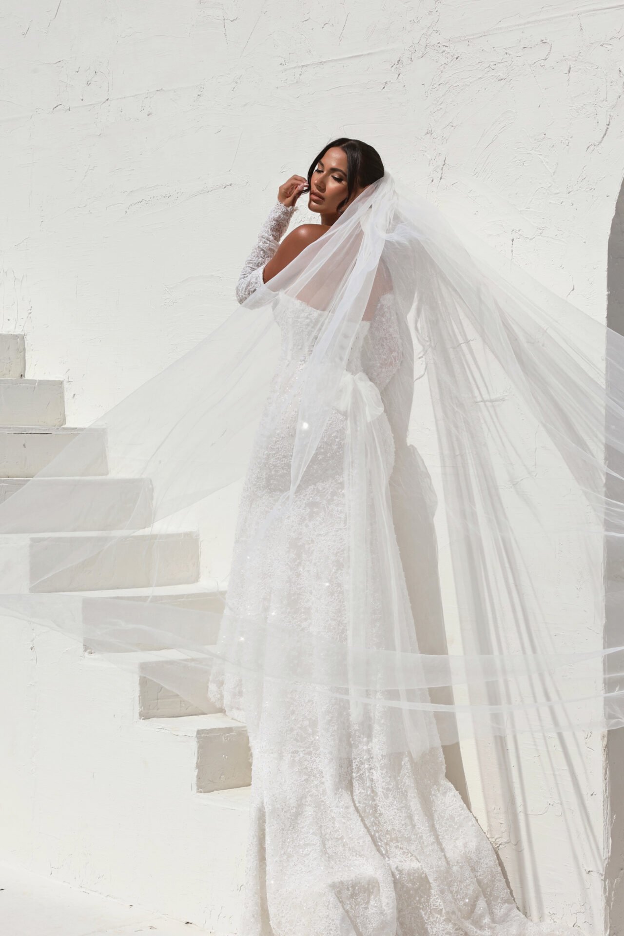 A woman wearing a wedding dress with a long veil posing on white stairs against a white textured wall.