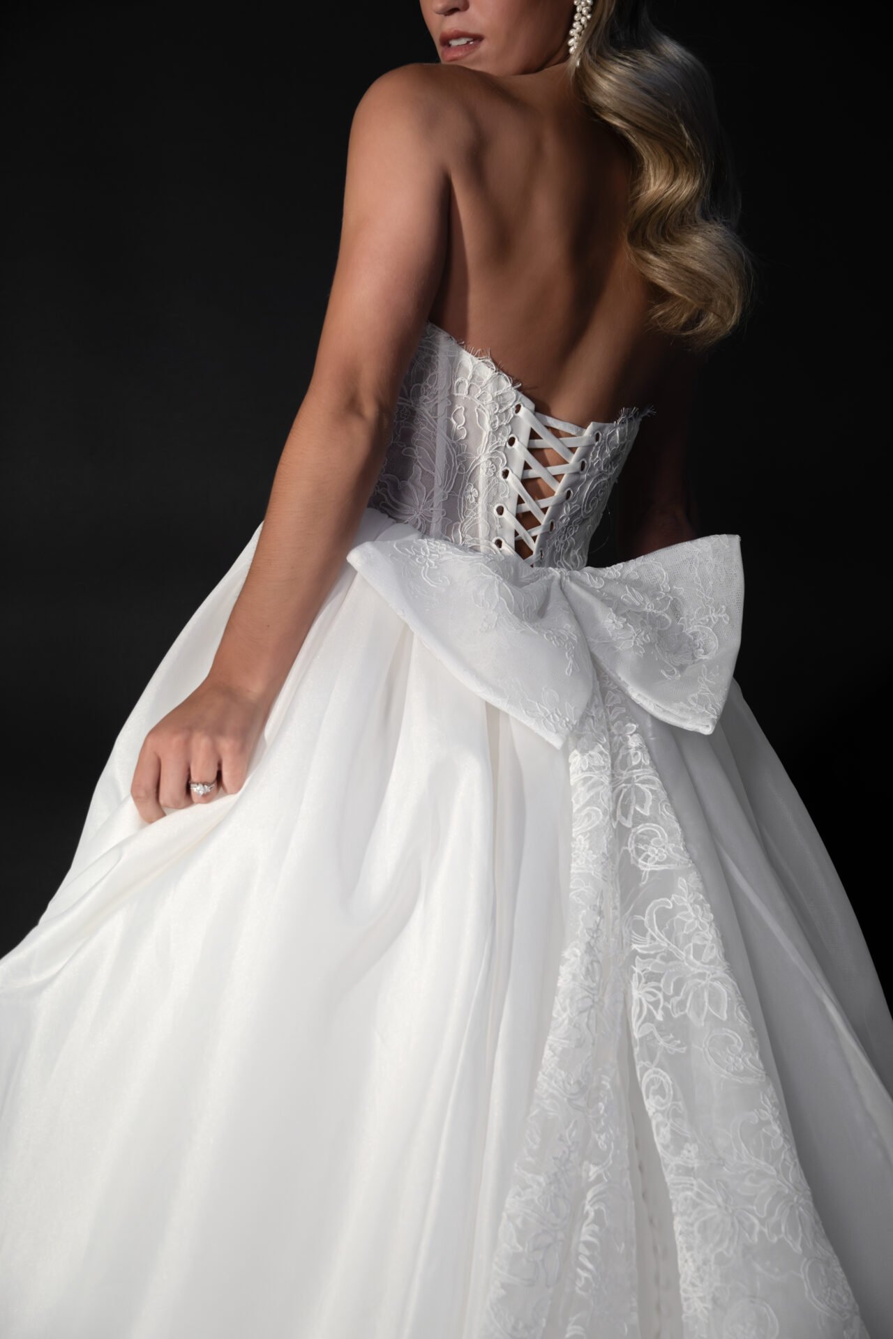 A woman dressed in an elegant white wedding gown with floral embroidery, a large bow, and a lace-up corset back, against a dark background.