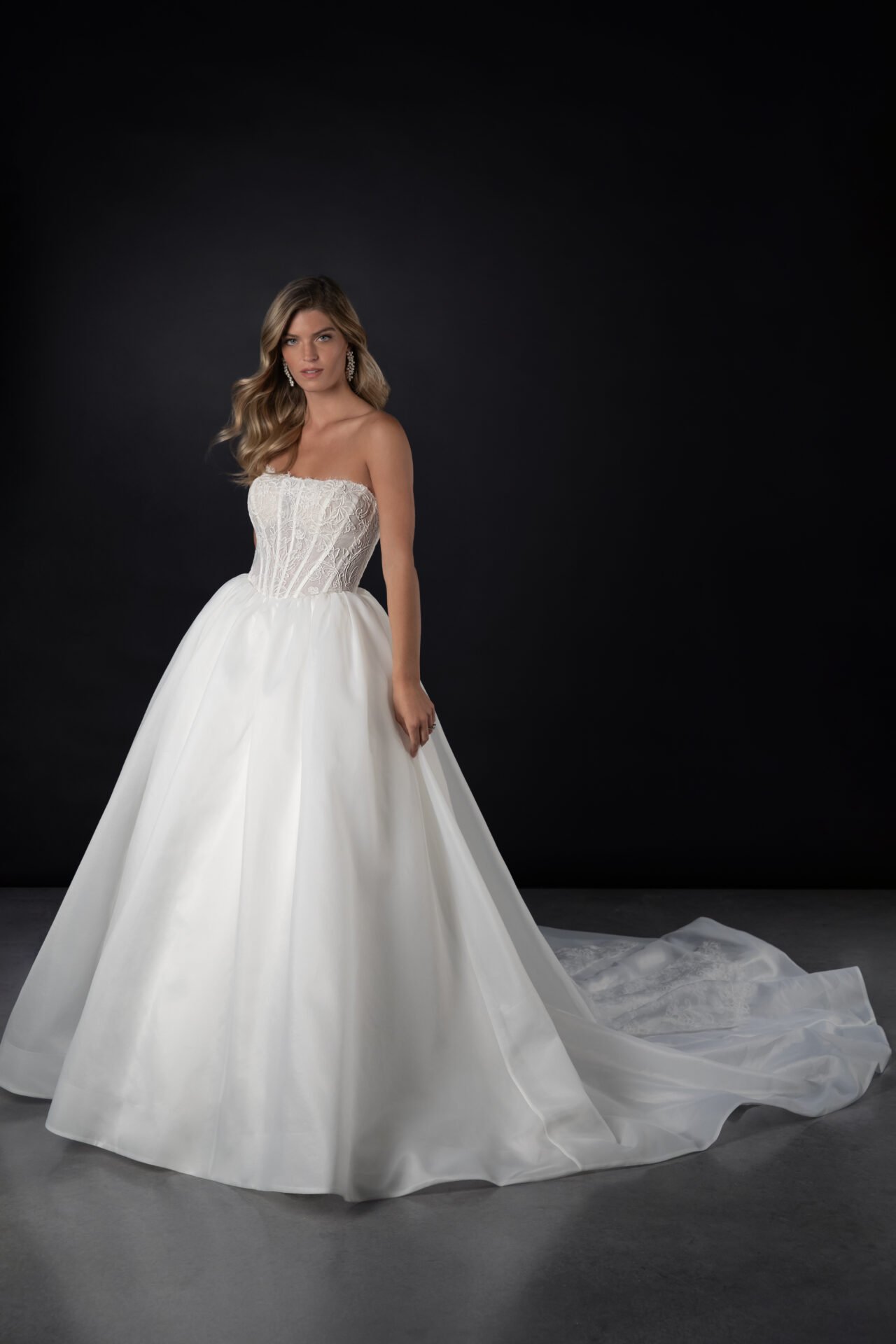 A woman wearing a strapless white wedding gown with a lace bodice and full skirt standing against a dark background.
