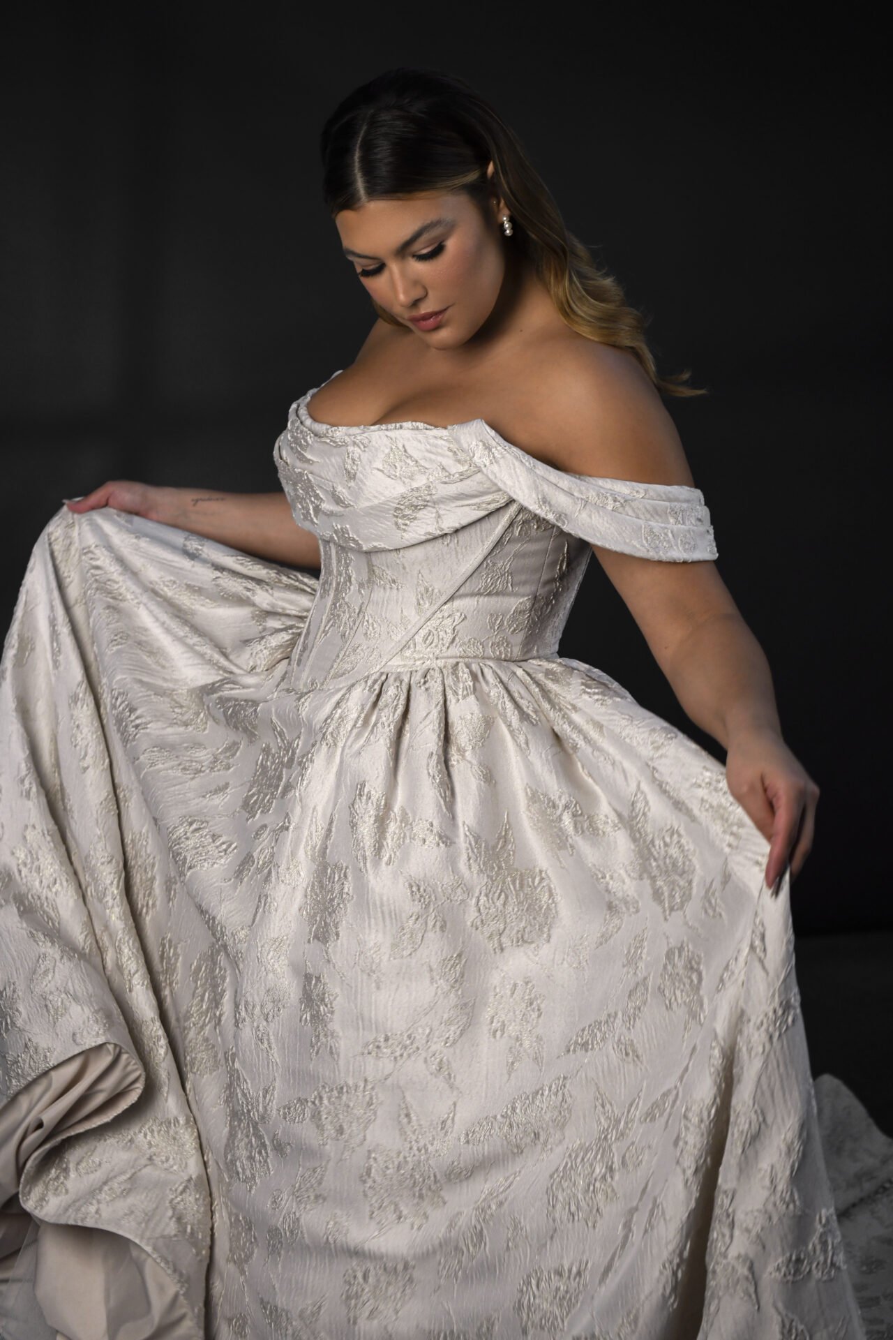 A woman wearing an off-the-shoulder, cream-colored, patterned ball gown, looking down and holding the skirt of her dress.