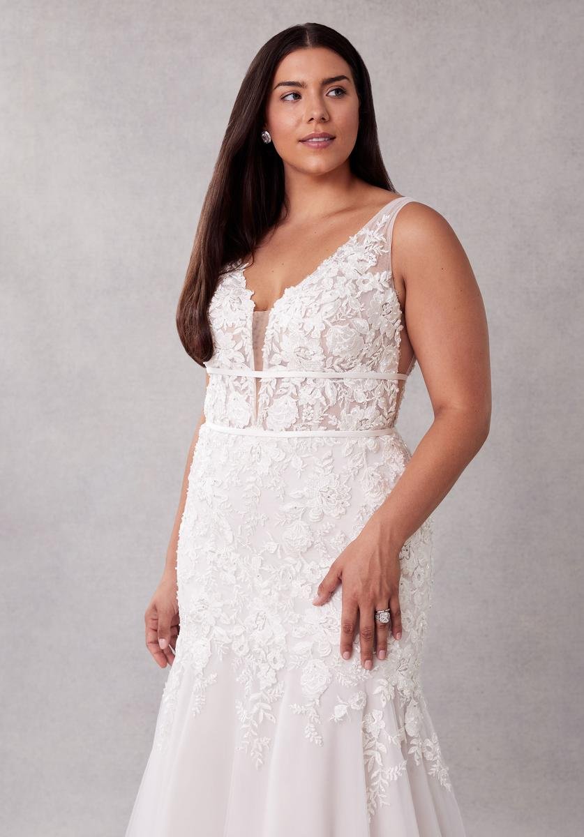 A woman with long dark hair wearing a white lace wedding dress with a deep V neckline and floral embroidery, standing against a neutral background.