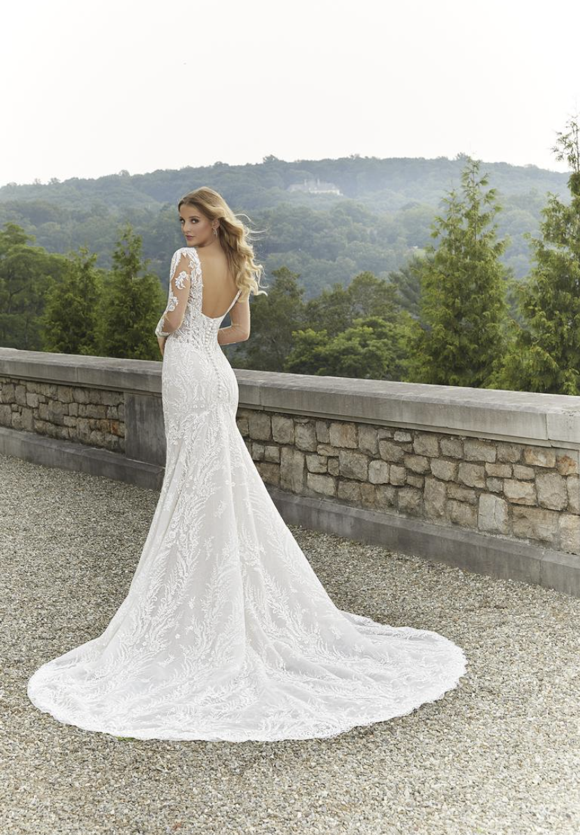 Bride in a white lace wedding gown standing outdoors on a gravel pathway, with a stone wall and green hills in the background.