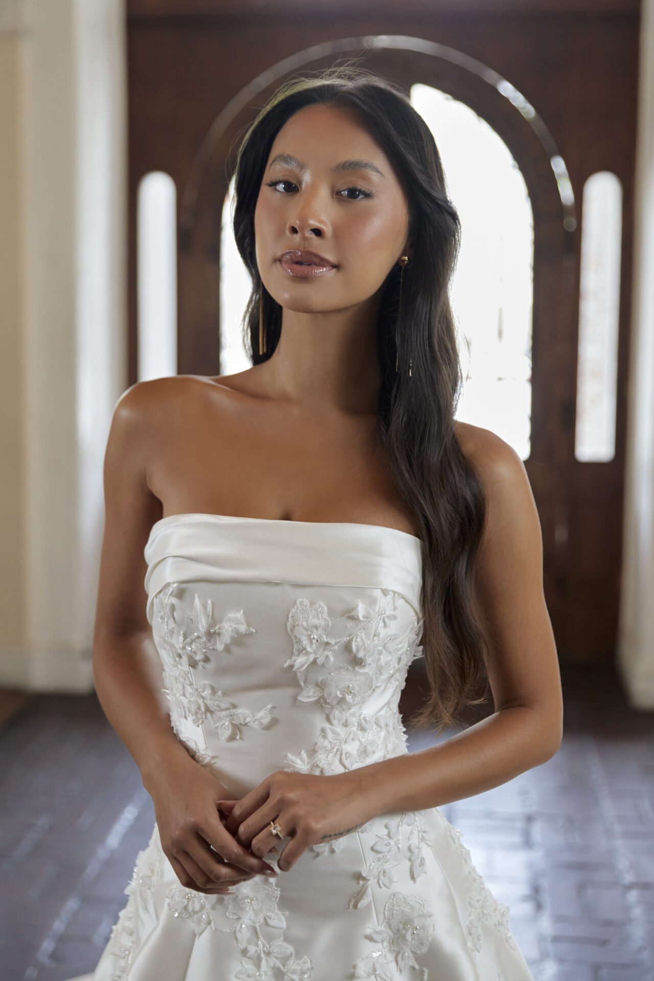 A woman wearing a strapless white wedding gown with floral embroidery, standing indoors near a wooden door with arched windows.