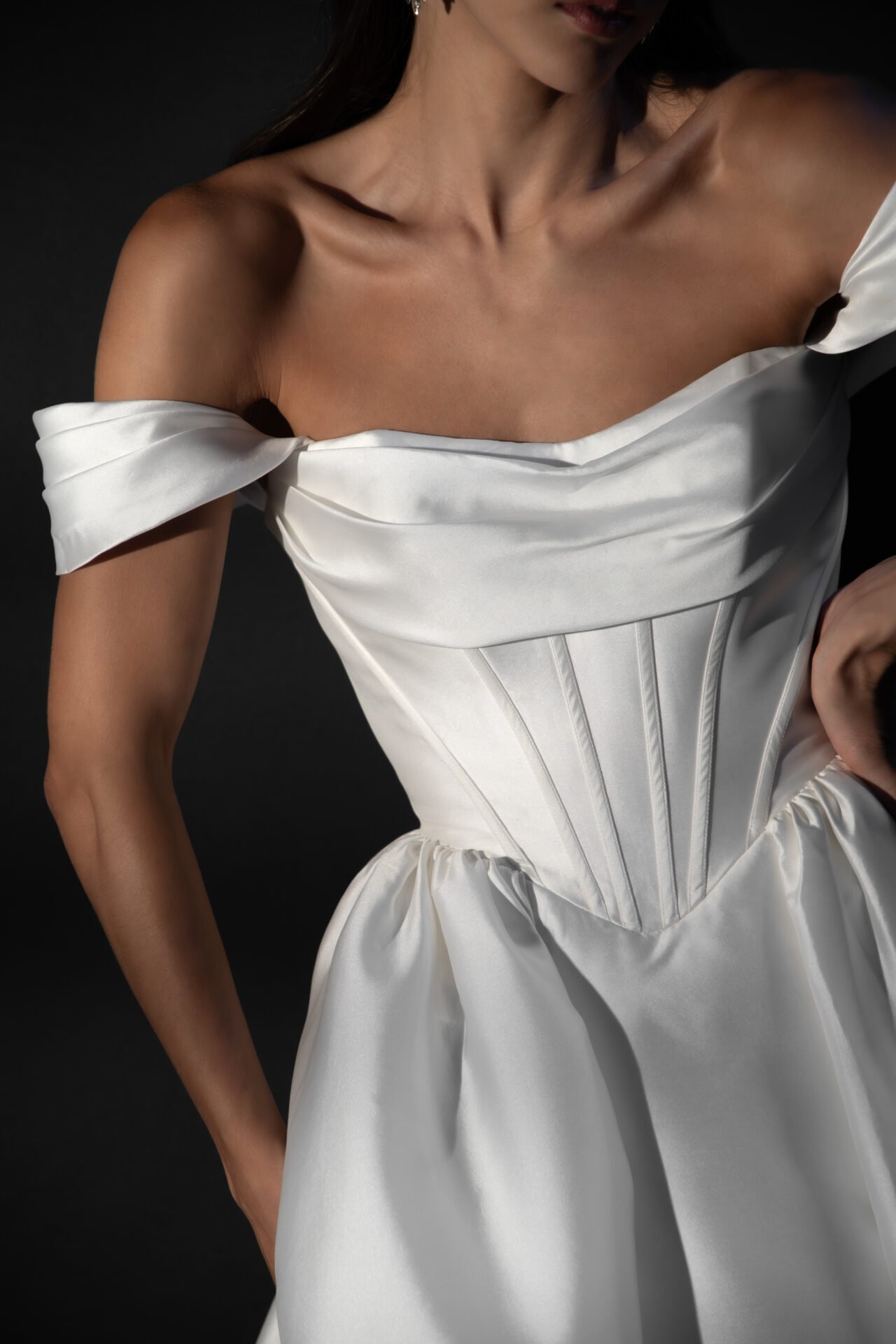 Close-up of a woman wearing a white satin off-shoulder dress with a fitted bodice and puffed sleeves against a black background.