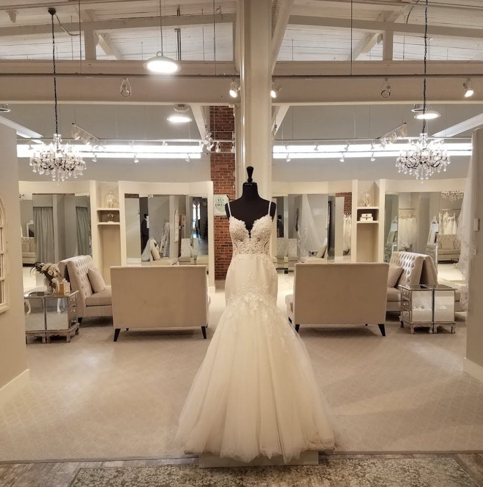 White wedding dress displayed on a mannequin in a bridal boutique with elegant chandeliers and seating area in the background.