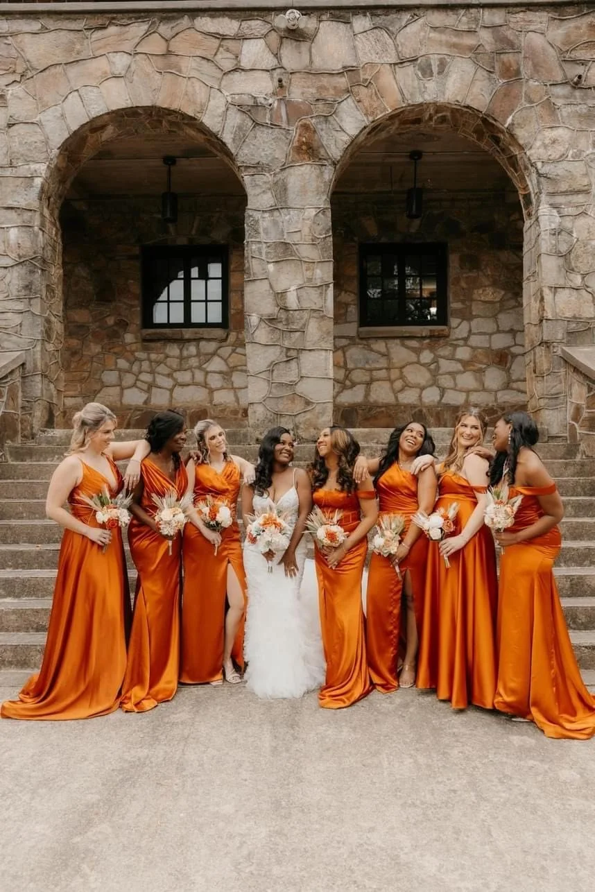 A bride and seven bridesmaids standing on stone steps in front of a stone building with two arched doorways, all dressed in orange dresses except the bride in a white wedding gown, holding bouquets of flowers, smiling and laughing.