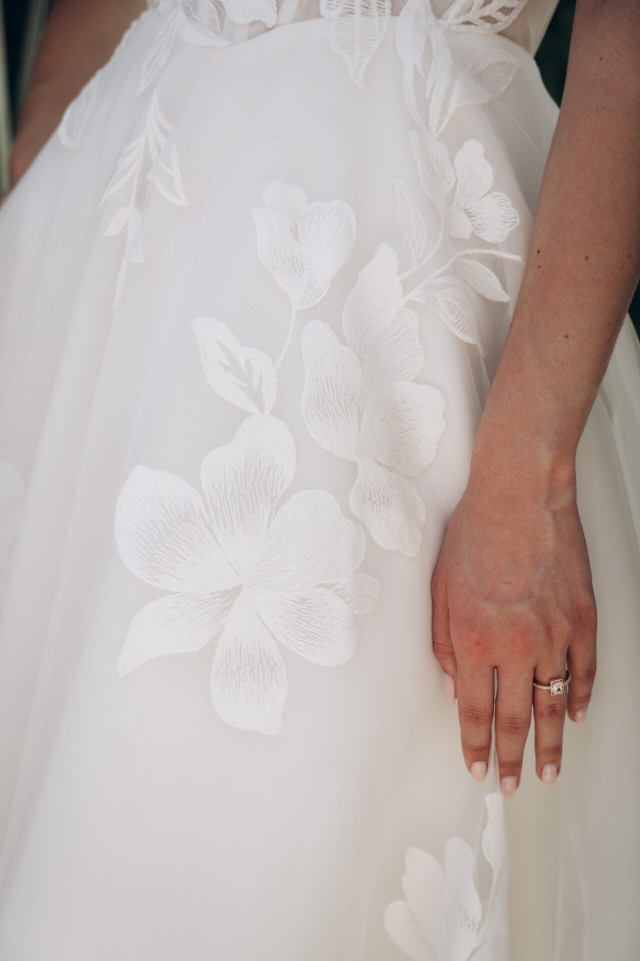 Close-up of a wedding dress with embroidered white floral patterns and a woman's hand with a ring resting on the fabric.