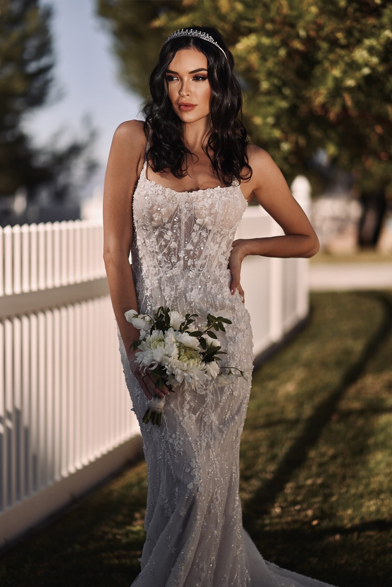 A woman in a white lace wedding gown with dark wavy hair and a tiara holding a bouquet of white flowers, standing outdoors with a white fence and trees in the background.