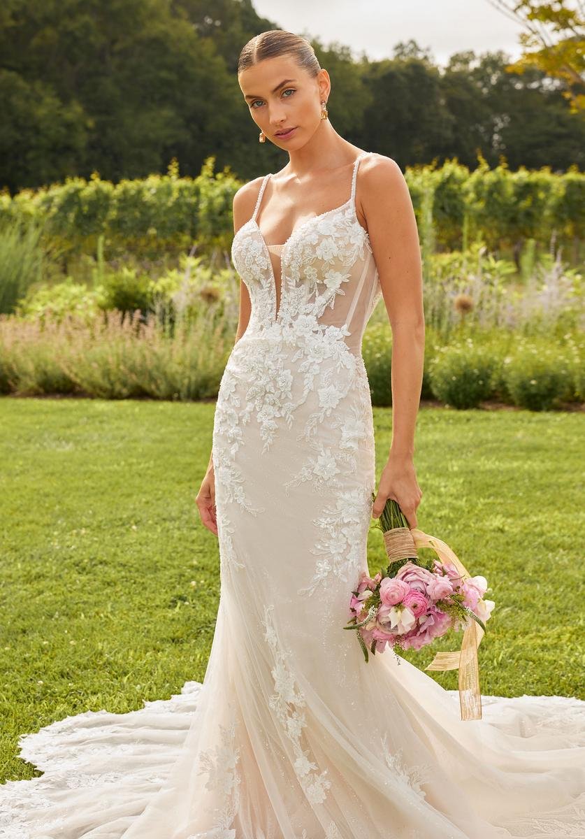 Bride in an elegant white lace wedding dress holding a bouquet of pink and white flowers standing on green grass in a garden setting.