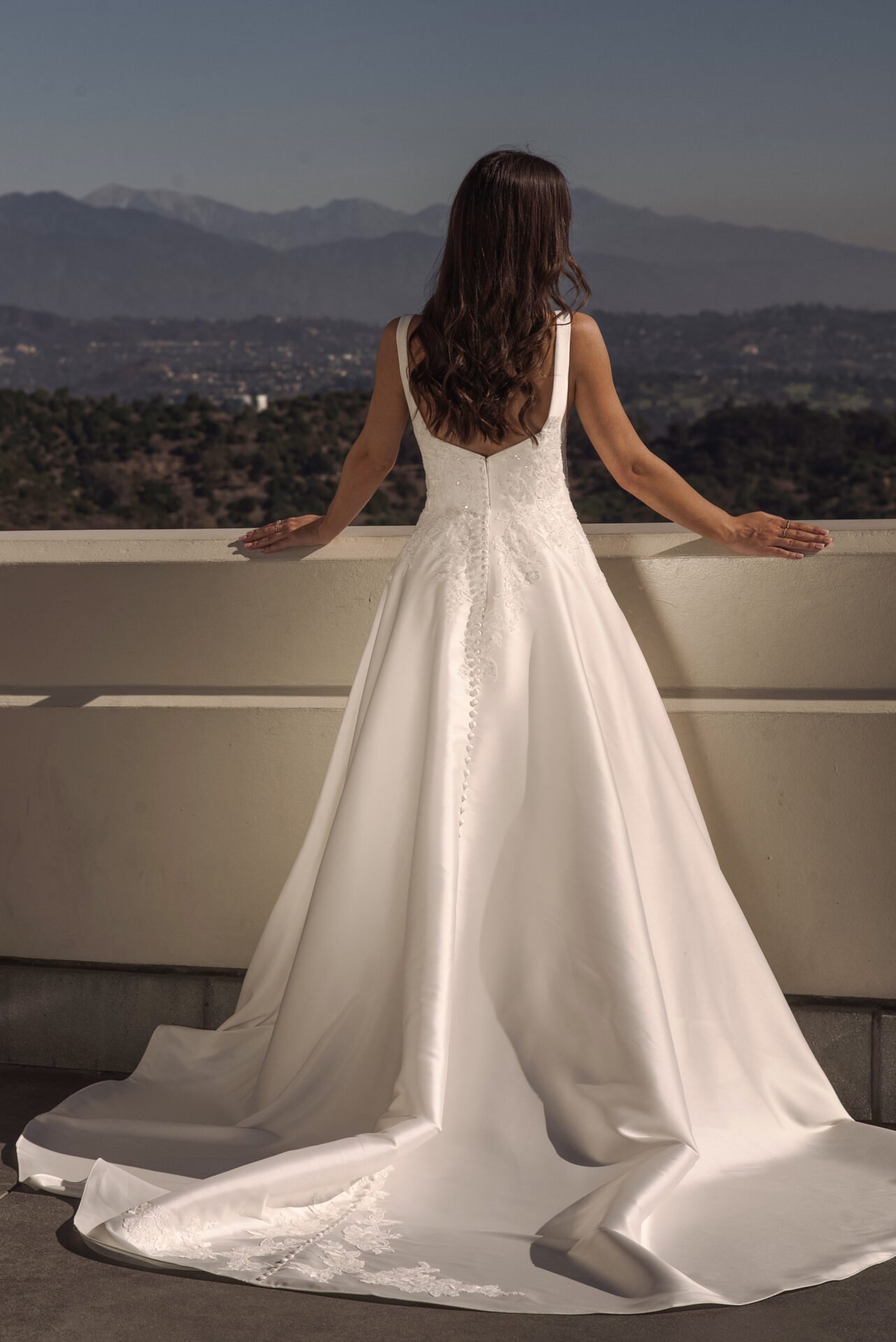 A woman in a wedding dress standing on a balcony, facing a mountainous landscape at dusk.