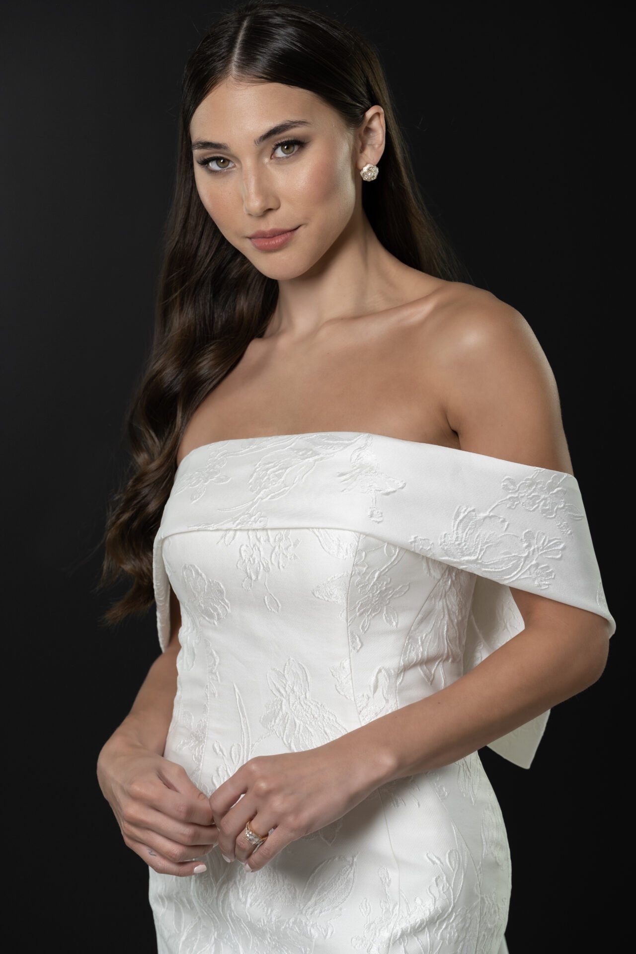 A woman in an off-the-shoulder white dress with floral embroidery, dark hair styled in loose waves, and wearing earrings and a ring, stands against a dark background.