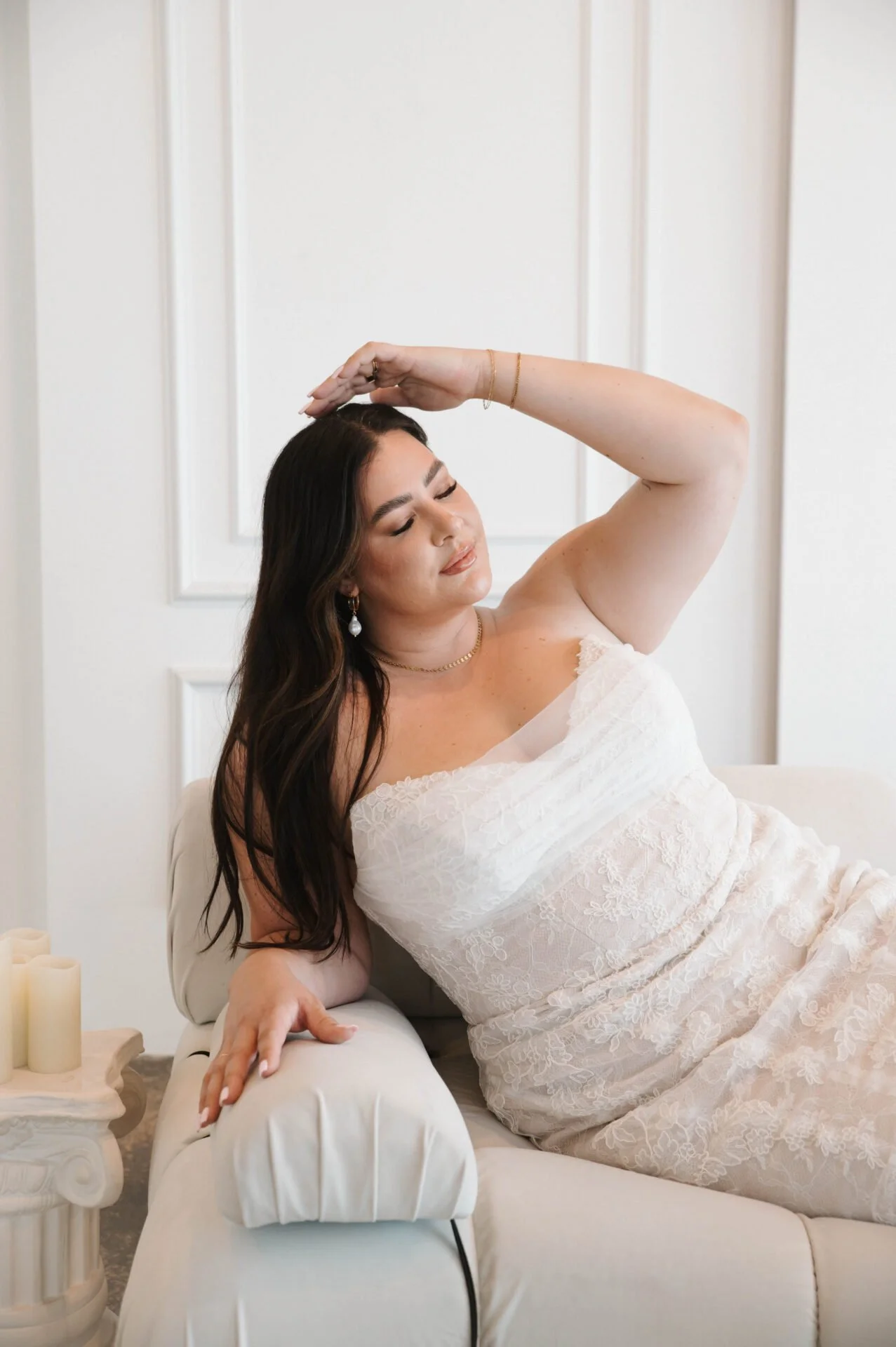 A woman in a white lace dress relaxing on a cream-colored sofa with her eyes closed, resting her head on her right hand raised above her head, in a bright room with white paneled walls and candles on a side table.