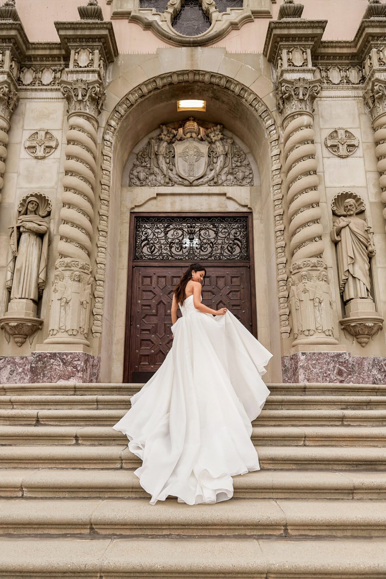 A woman in a white wedding dress standing on the steps of a church with a detailed, ornate stone facade and statues.
