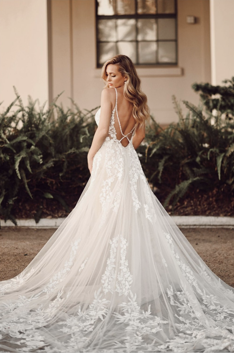 A woman in a white wedding gown with lace details and an open back, standing outdoors on a dirt path, with green bushes and a building in the background.
