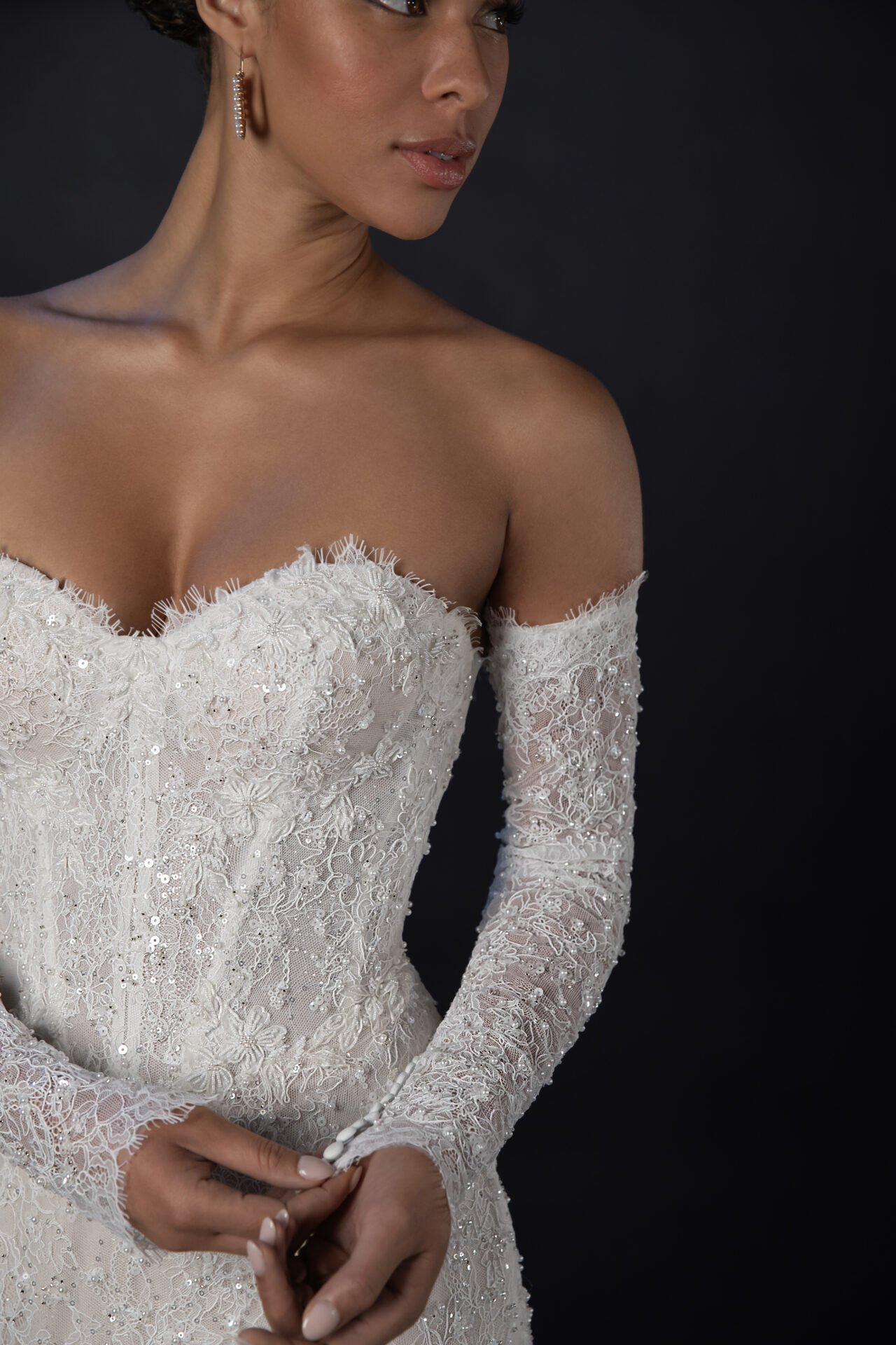 Close-up of a woman in a white lace wedding dress with long sleeves, against a dark background.
