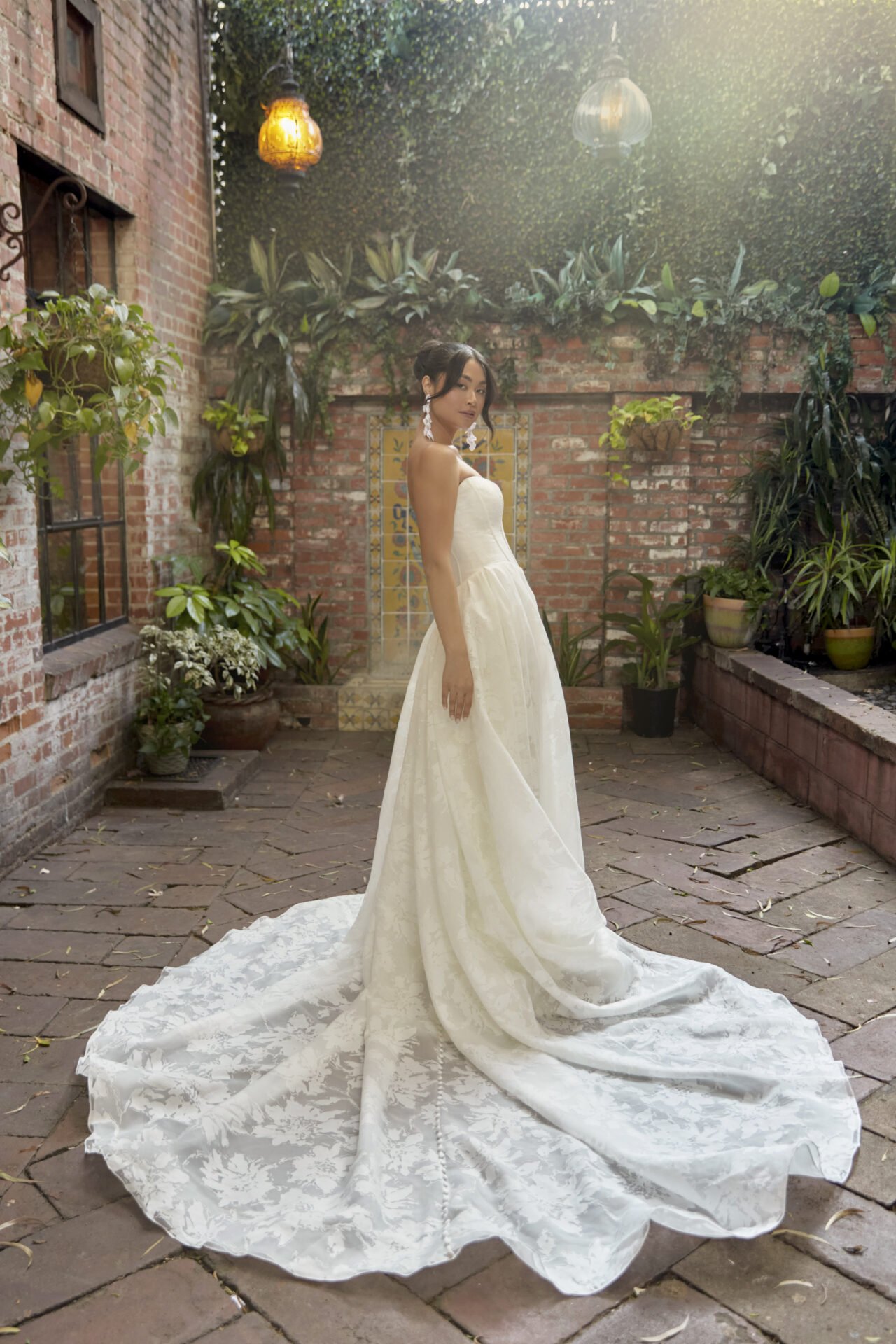 A woman in a strapless white wedding dress with a long train stands in a brick courtyard surrounded by green plants and hanging lanterns.