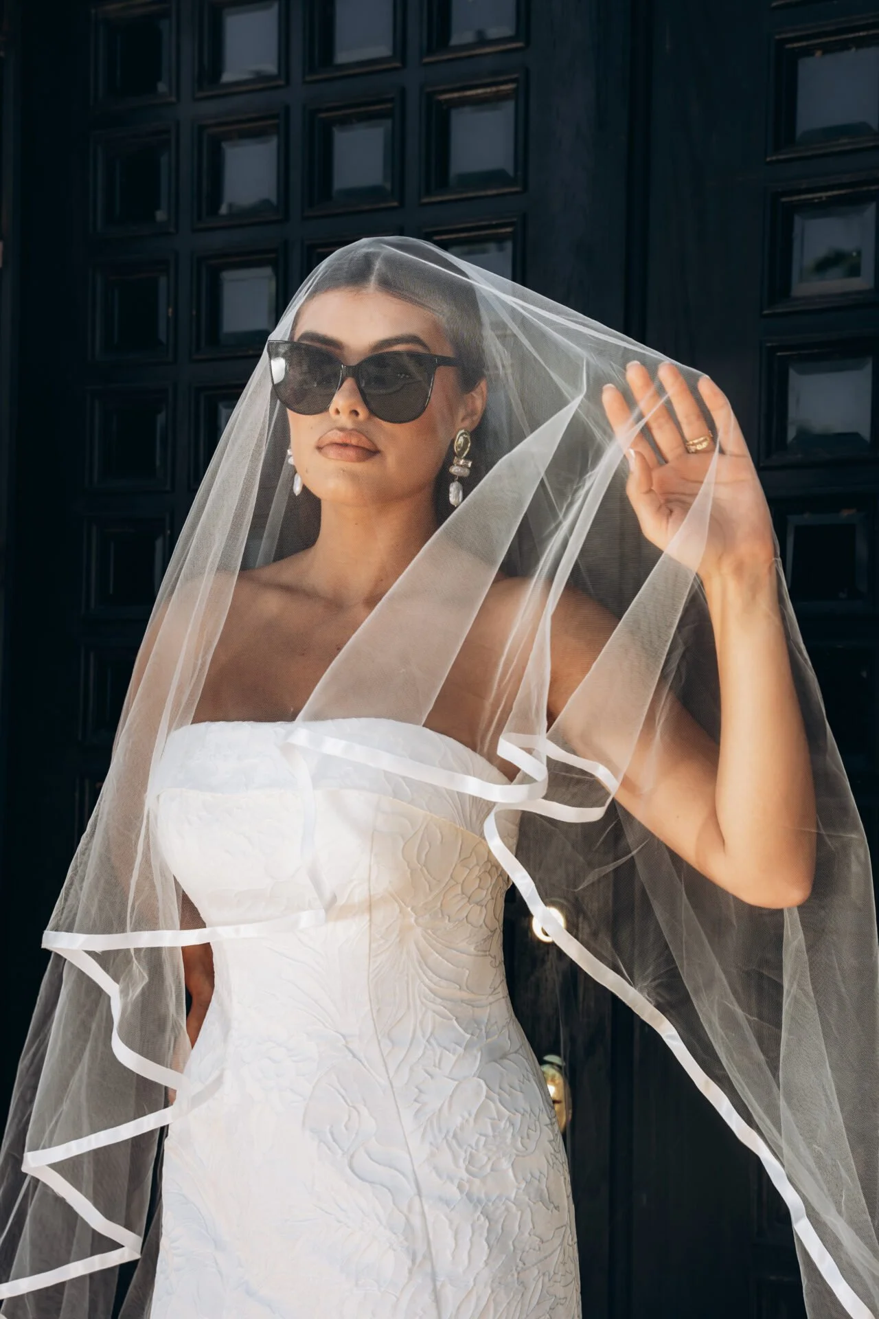 A woman in a white wedding dress accessorized with large earrings and dark sunglasses, holding a sheer veil over her head. She is standing in front of a black wooden door.