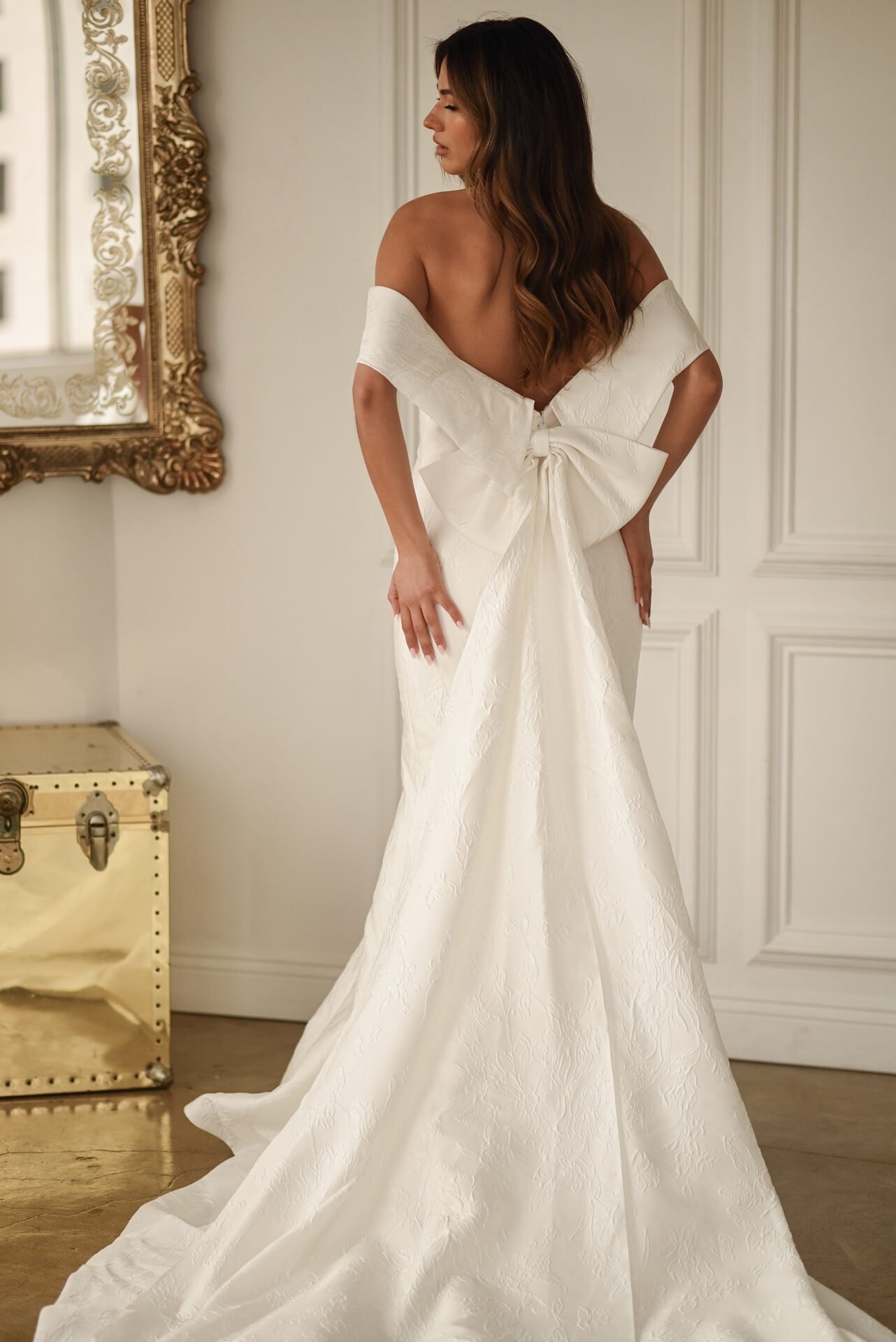 Woman in an elegant white wedding gown with a large bow on the back, standing indoors with her back to the camera, near a decorative mirror and a vintage chest.
