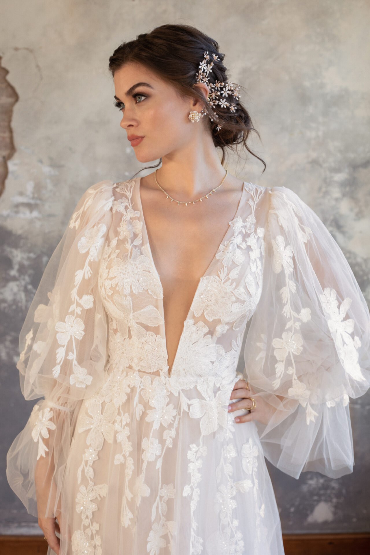 A woman in a white, floral lace wedding gown with puffed sleeves, wearing wedding jewelry, with an ornate hairpiece, standing against a textured wall.
