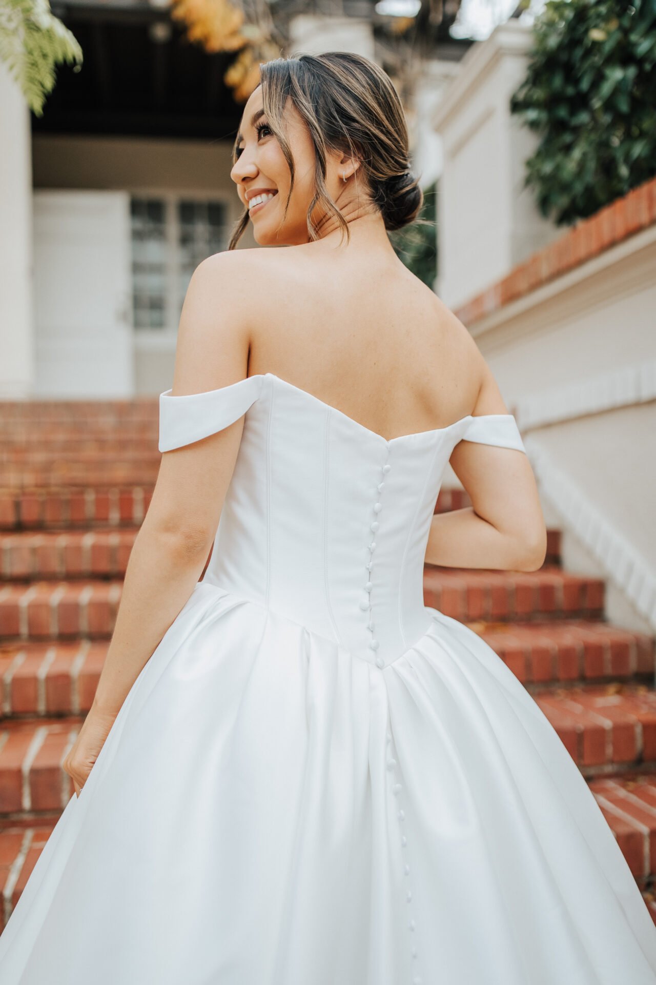 A smiling woman with dark hair in an elegant white off-the-shoulder wedding gown with buttons down the back, standing on brick stairs outside a house.