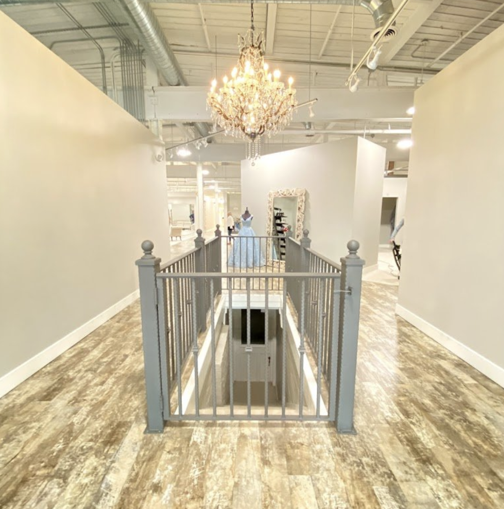 Interior of a retail space with a chandelier, mannequin with a blue dress, large mirror, and staircase leading downstairs.