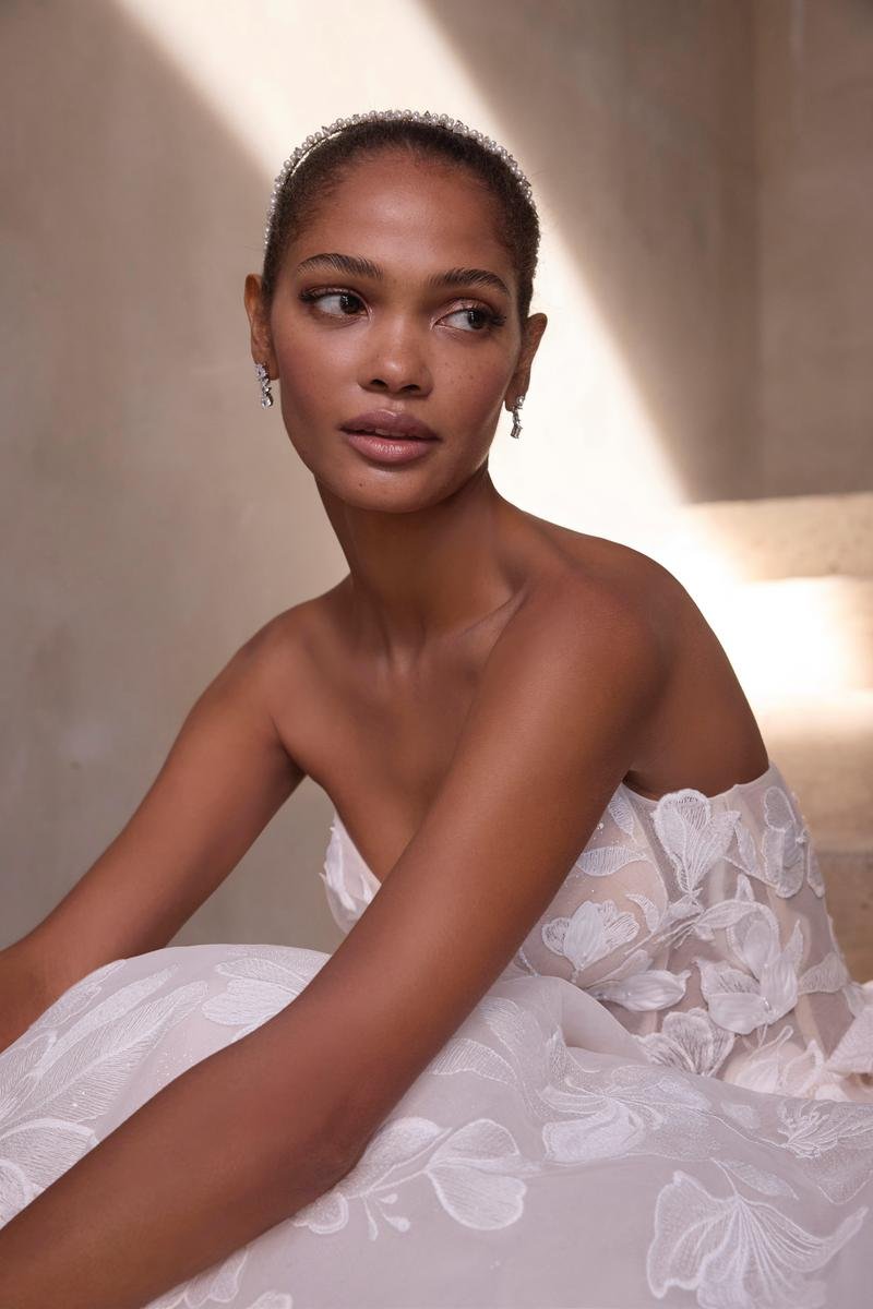 A woman in a white lace dress and earrings, wearing a tiara, sitting indoors with soft lighting.